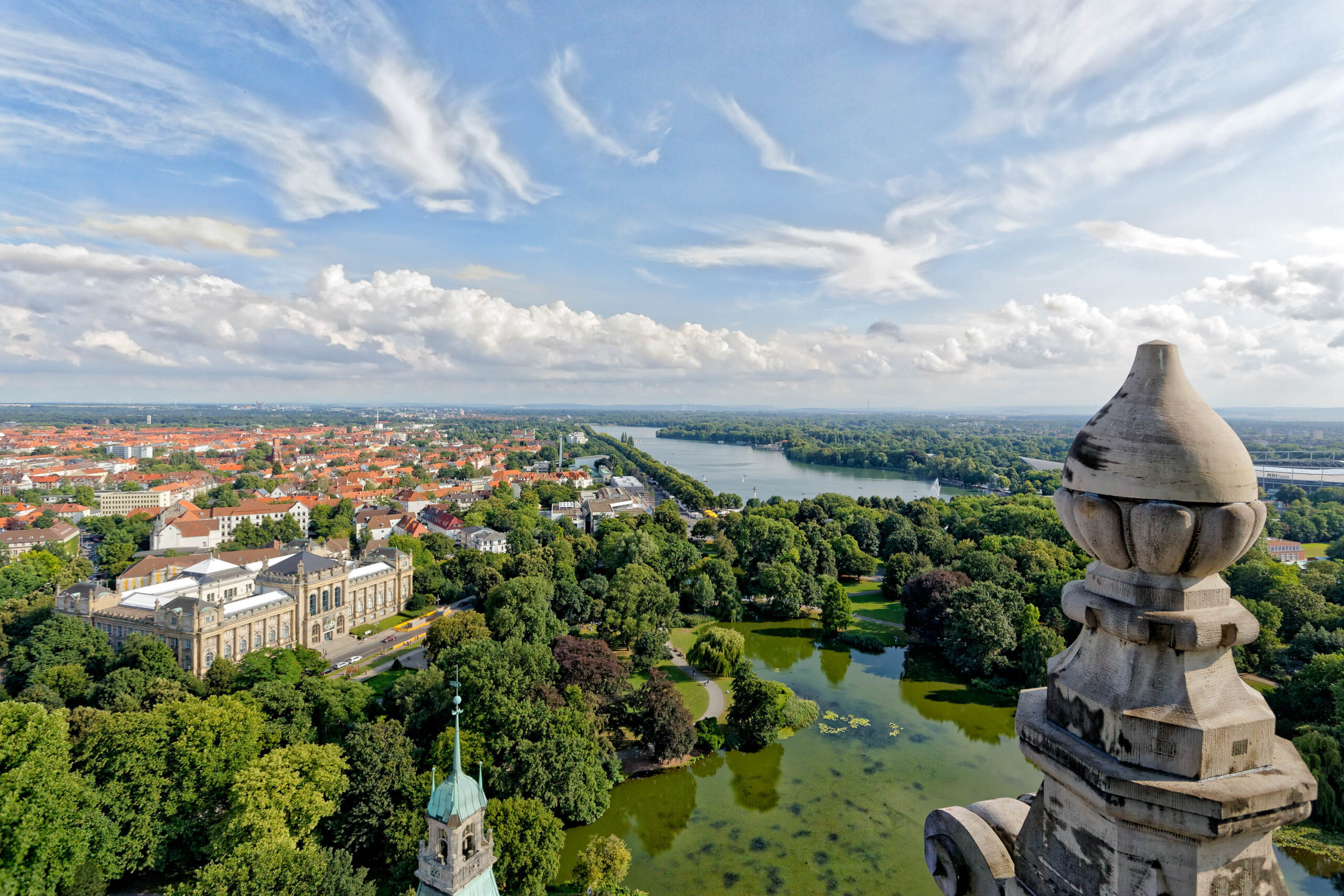 Ein atemberaubender Panoramablick auf die Stadt Hannover, aufgenommen aus einer erhöhten Perspektive, vermutlich von einem Kirchturm oder einem ähnlichen erhöhten Punkt. Im Vordergrund erstreckt sich ein üppiger, grüner Park, der zum Maschsee übergeht, einem See, der von Bäumen umgeben ist. Im Hintergrund erhebt sich die Stadtlandschaft mit ihren roten Ziegeldächern und historischen Gebäuden. Das Schloss von Hannover ist deutlich erkennbar und prägt das Stadtbild. Der Himmel ist blau mit vereinzelten weißen Wolken, was auf einen sonnigen Tag hindeutet. Die Szene vermittelt ein Gefühl von Ruhe, Weite und historischer Bedeutung.