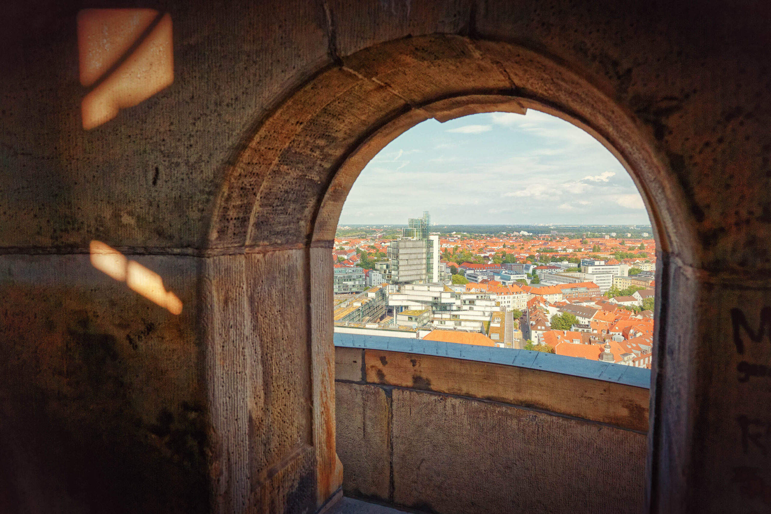 Das Bild zeigt eine Perspektive durch ein Rundbogenfenster, das einen Blick auf die Stadt Hannover freigibt. Das Fenster selbst ist aus dunklem Stein oder Beton gefertigt und wirkt rustikal und alt. Das Licht fällt schräg durch das Fenster und erzeugt interessante Licht- und Schattenspiele an den Innenwänden.  Die Stadt Hannover erstreckt sich im Hintergrund, mit einer Mischung aus roten Ziegeldächern und modernen Hochhäusern. Der Himmel ist leicht bewölkt, was für eine sanfte Beleuchtung sorgt. Die Szene vermittelt ein Gefühl von Ruhe und Kontemplation, da der Betrachter einen Blick auf die Stadt aus einer erhöhten Position erhält.