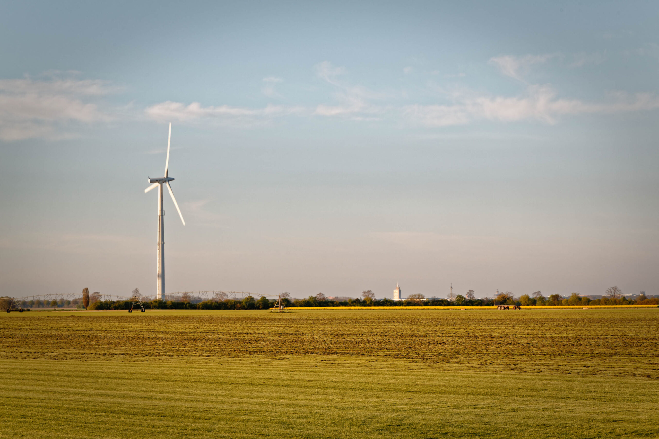 Die Aufnahme zeigt eine weite Landschaftsaufnahme, die ein Feld dominiert. Das Feld ist mit einer hellbraunen Erntefläche bedeckt, die im Vordergrund liegt. Im Hintergrund erheben sich mehrere Windkraftanlagen, die den Horizont dominieren. Der Himmel ist leicht bewölkt und hat einen bläulichen Farbton. Die Szene vermittelt ein Gefühl von Weite und Ruhe. Die Aufnahme wurde wahrscheinlich in der Region Calenberger Land, möglicherweise in Pattensen, aufgenommen.