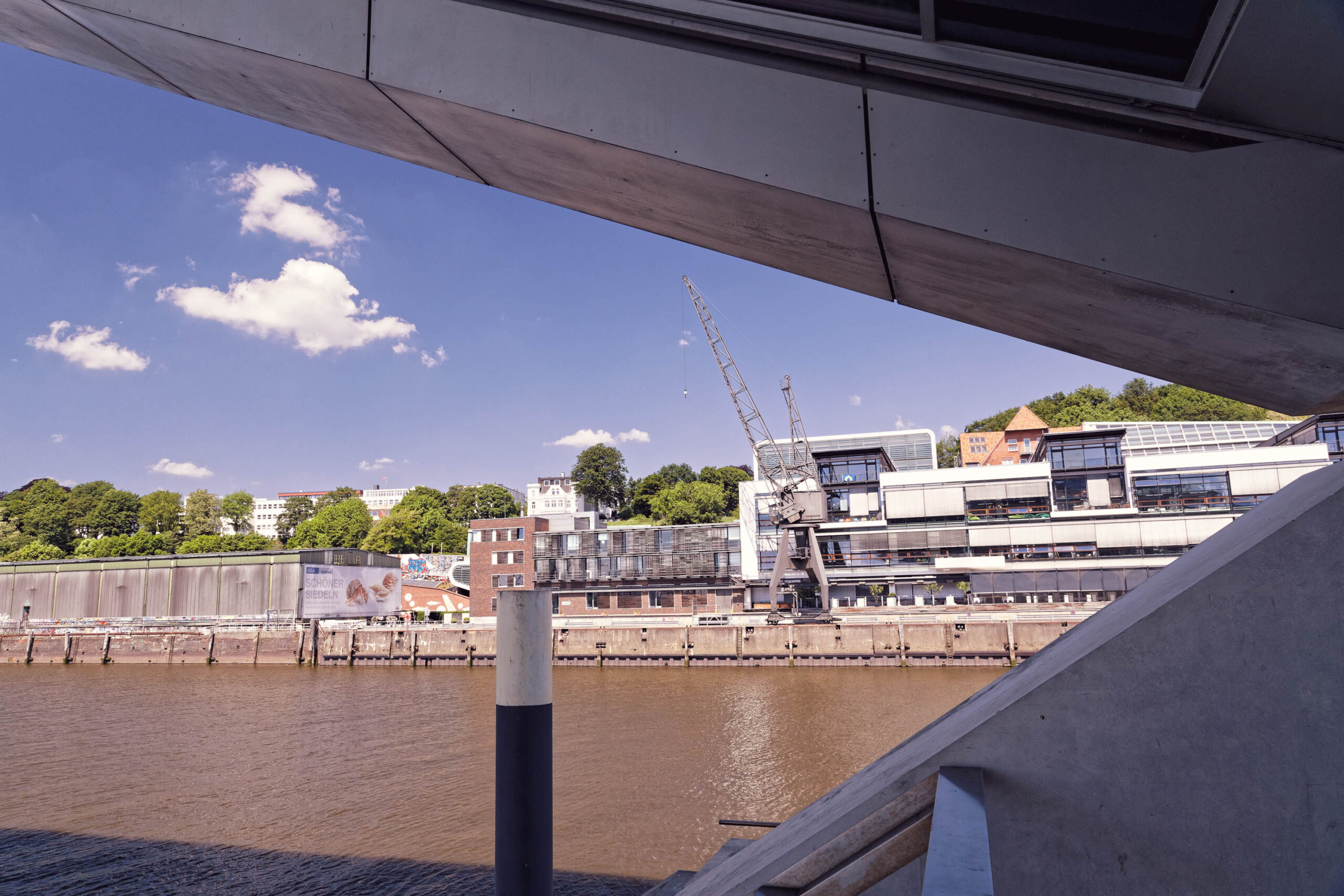 Die Aufnahme zeigt eine Perspektive von der Elbphilharmonie in Hamburg, aufgenommen im Mai 2018. Der Blick fällt auf die Elbe und die gegenüberliegenden Gebäude, die einen Teil der Hamburger Skyline bilden. Ein Kran deutet auf Bauarbeiten hin. Die Elbphilharmonie selbst ist teilweise durch die Brüstung des Geländers verdeckt. Der Himmel ist blau und von vereinzelten weißen Wolken durchzogen. Die Szene vermittelt eine ruhige und sonnige Atmosphäre.