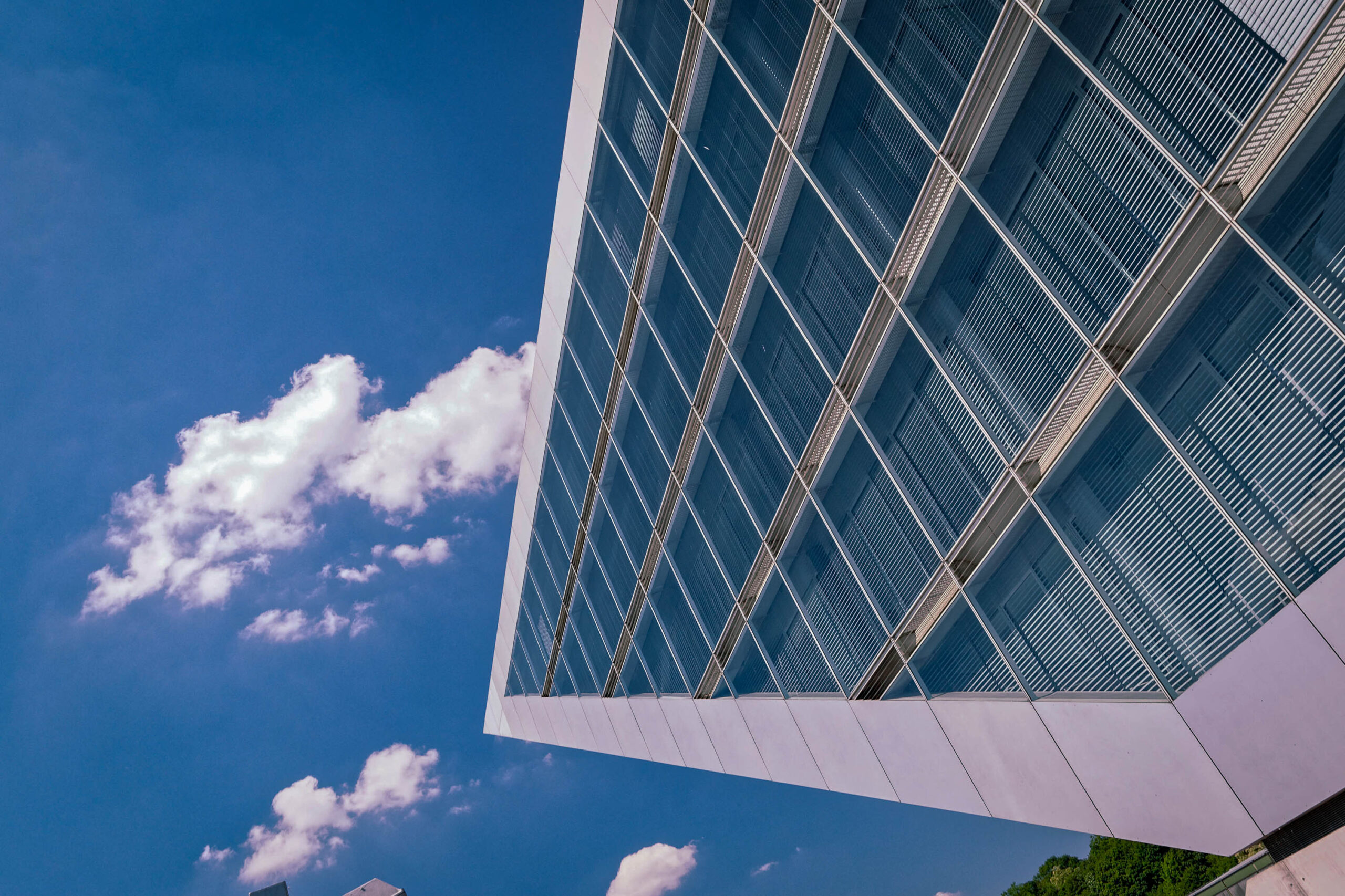 Das Foto zeigt eine ungewöhnliche Perspektive auf ein modernes Bürogebäude in Hamburg. Der Blickwinkel ist nach oben gerichtet, wodurch das Gebäude dominant im Bild erscheint. Die Fassade besteht aus einer Kombination aus Glas und weißen Paneelen, die das Sonnenlicht reflektieren. Der Himmel ist blau und von weißen, flauschigen Wolken bedeckt. Die Komposition betont die Höhe und die architektonische Gestaltung des Gebäudes. Die Aufnahme wurde im Mai 2018 aufgenommen.