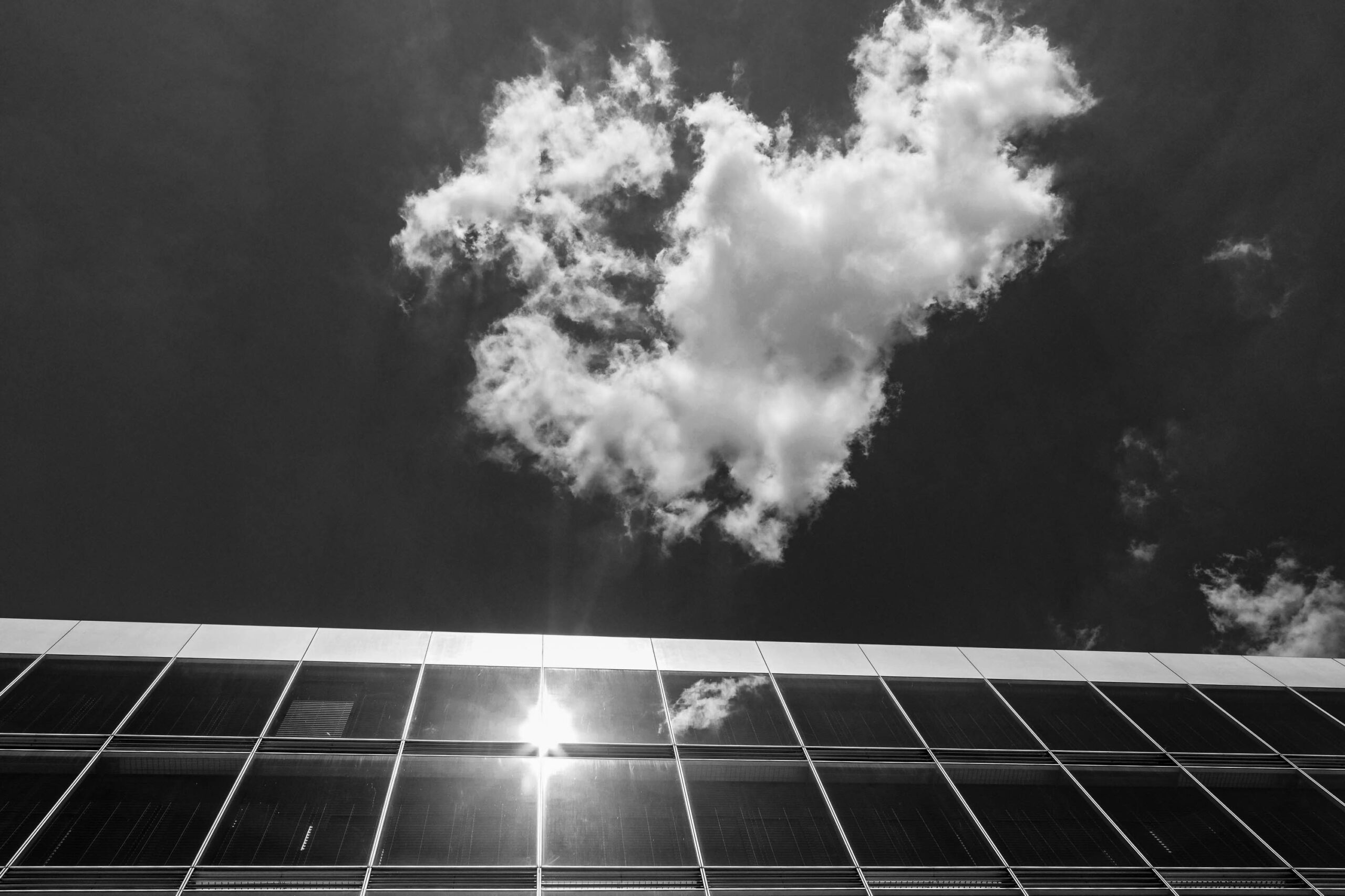 Das Foto zeigt eine moderne Gebäudefassade in Schwarzweiß. Die Fassade besteht aus einer großen Fläche von quadratischen Fenstern, die das Sonnenlicht reflektieren. Der Himmel darüber ist von dichten, weißen Wolken bedeckt. Die Komposition betont die geometrische Struktur des Gebäudes im Kontrast zum organischen Muster der Wolken. Das Foto wurde im Mai 2018 in Hamburg aufgenommen.