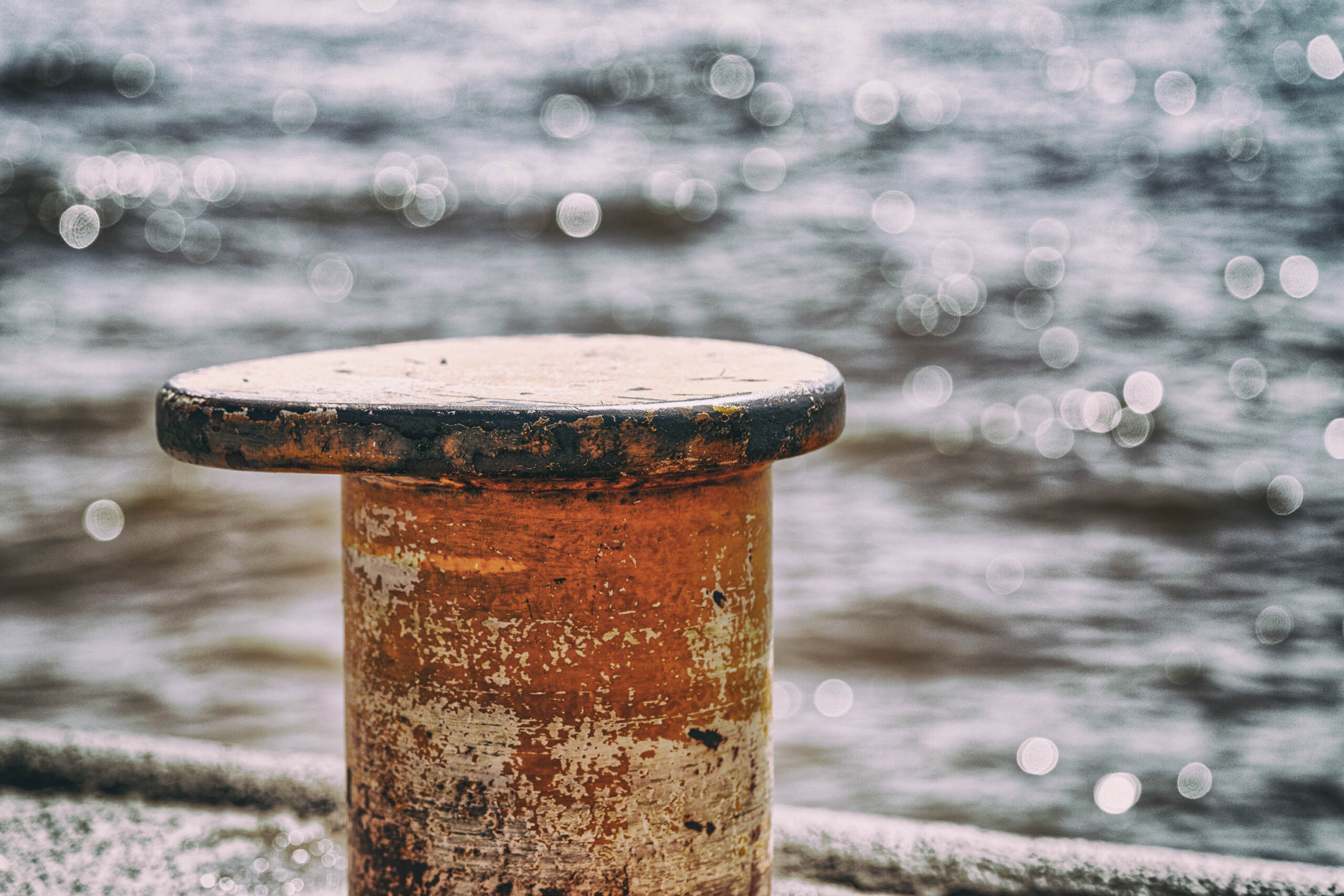 Das Bild zeigt eine Nahaufnahme eines alten, rostigen Ankers. Der Anker ist hauptsächlich orangefarben, mit schwarzen Akzenten und deutlichen Rostflecken. Die Textur des Metalls ist sichtbar, mit Abnutzungserscheinungen und Verwitterung. Der Hintergrund ist unscharf und zeigt eine Wasseroberfläche, was darauf hindeutet, dass sich der Anker in der Nähe eines Hafens oder einer Küste steht. Die Beleuchtung ist gedämpft, was eine melancholische Atmosphäre erzeugt. Der Fokus liegt auf dem Anker, der die Geschichte und die raue Schönheit des Meeres widerspiegelt.