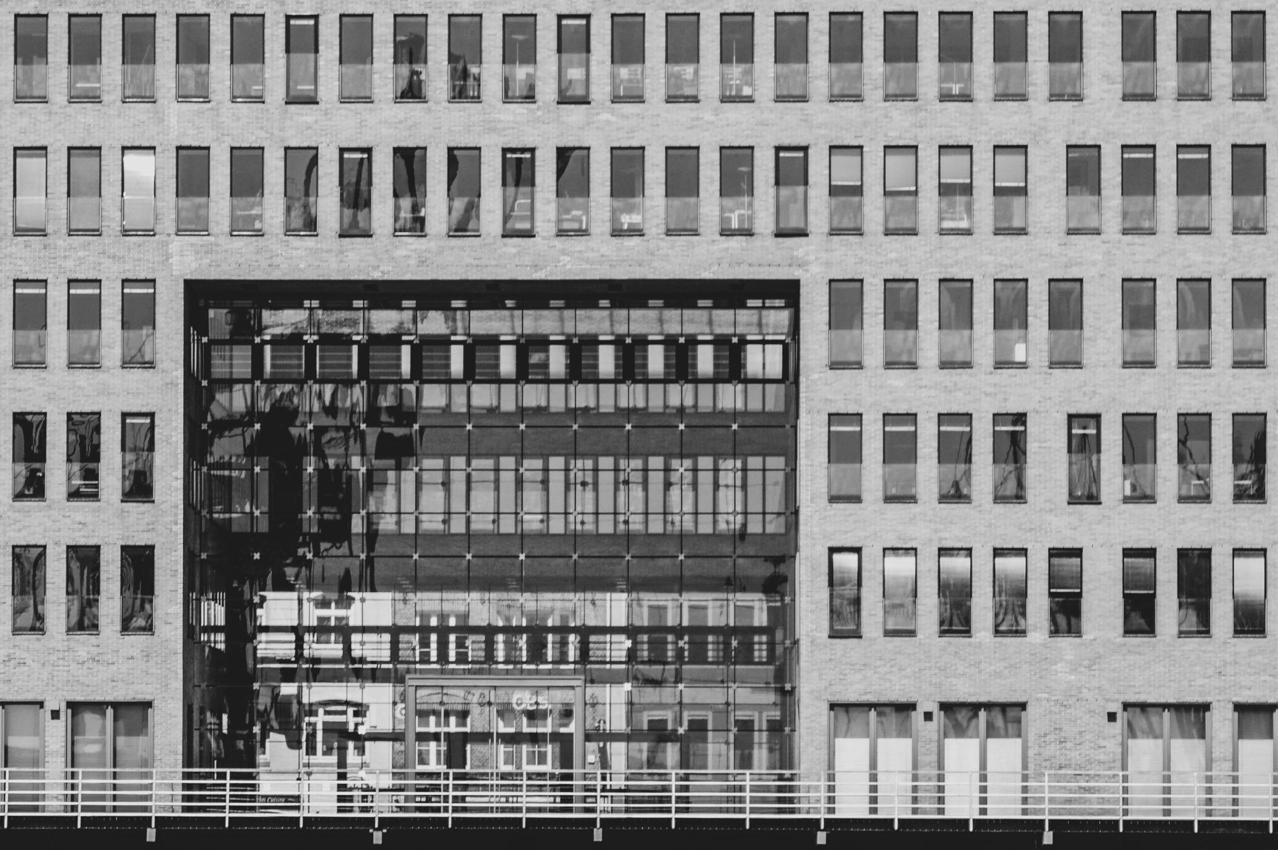 Das Foto zeigt die Fassade eines Bürogebäudes in Hamburg, aufgenommen im Mai 2018. Die Aufnahme ist in Schwarzweiß gehalten und betont die geometrische Struktur der Fassade. Das Gebäude besteht aus einer Vielzahl von Fenstern, die in einem Raster angeordnet sind. Ein großer, rechteckiger Bereich in der Mitte der Fassade ist durch eine Glaswand freigelegt, die den Innenraum des Gebäudes teilweise sichtbar macht. Die Fassade besteht aus hellem Backstein oder Beton, der im Kontrast zu den dunklen Fensterrahmen steht. Die Aufnahme fängt die architektonische Einfachheit und die repetitive Natur des Gebäudes ein. Die Beleuchtung erzeugt interessante Schatten und Lichtspiele auf der Fassade, die die Textur des Materials hervorheben. Die Aufnahme vermittelt einen Eindruck von Urbanität und Moderne.