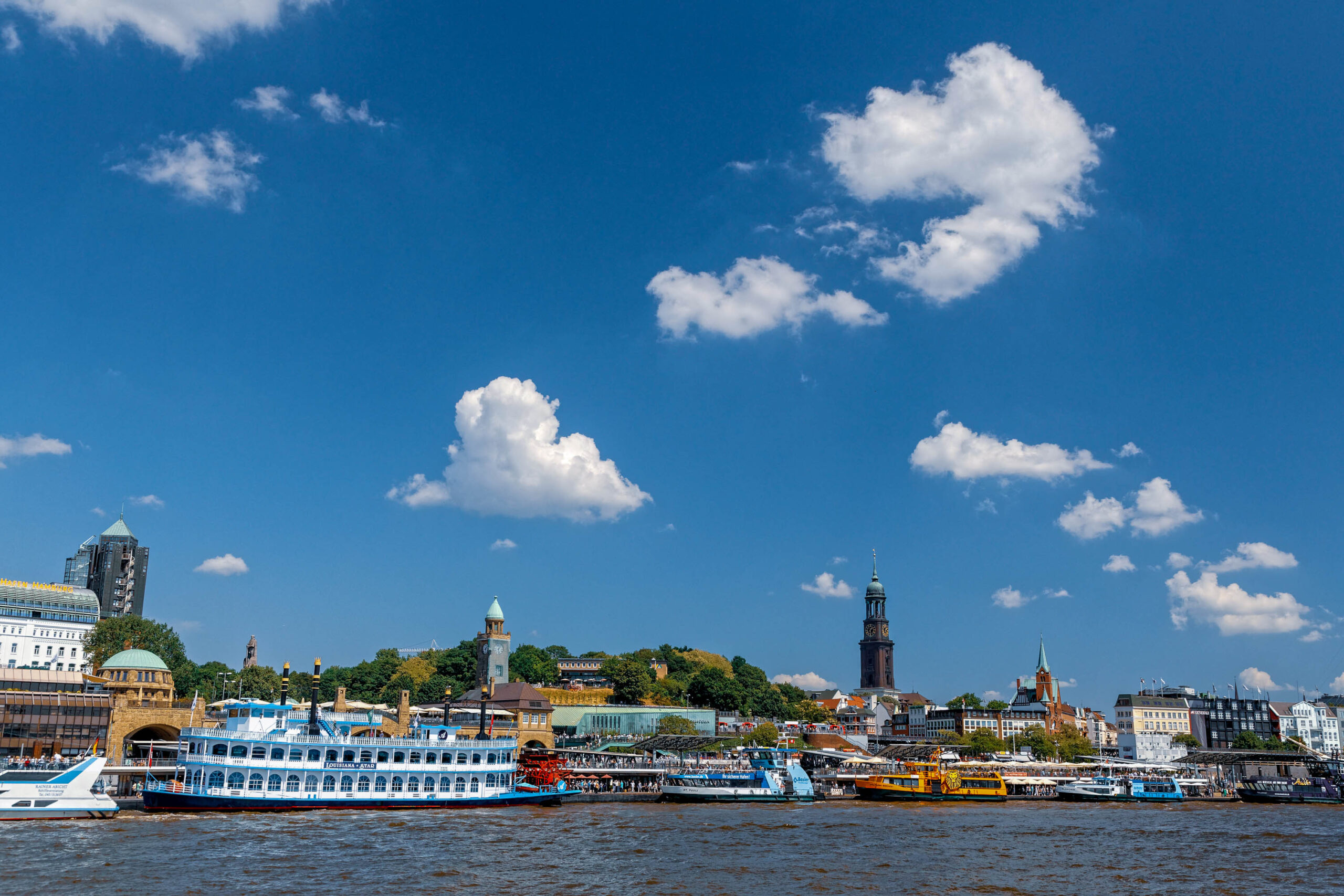 Die Aufnahme zeigt eine malerische Panoramaaufnahme von Hamburg, aufgenommen vom Wasser aus. Die Skyline der Stadt ist deutlich erkennbar, mit einer Mischung aus historischen und modernen Gebäuden. Der Himmel ist strahlend blau mit vereinzelten weißen Wolken, was eine sommerliche Atmosphäre schafft. Im Vordergrund befindet sich ein Fluss, der von Schiffen und Booten belebt wird. Die Komposition betont die Schönheit und den Charme der Stadt Hamburg.