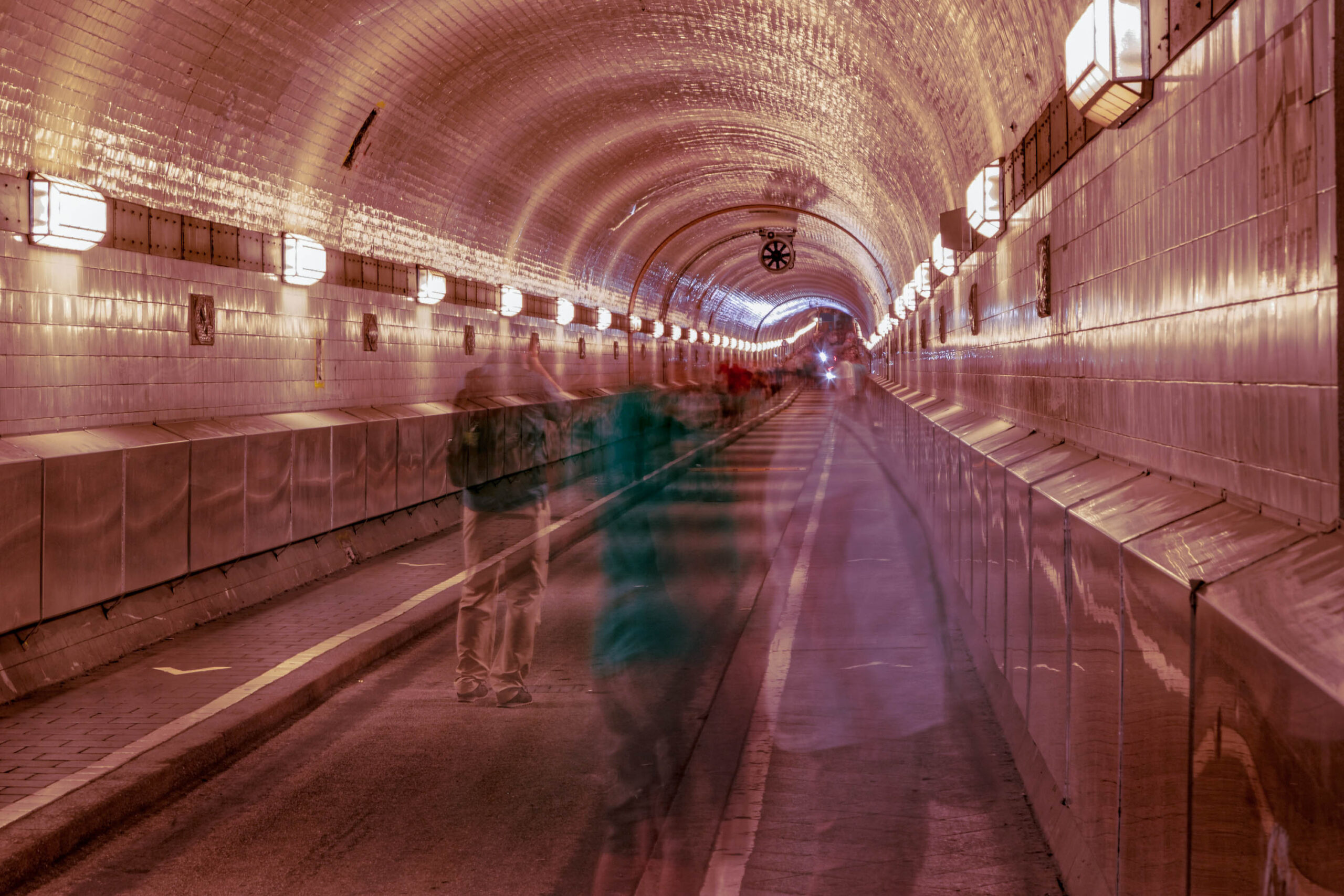 Die Aufnahme zeigt den St. Pauli Tunnel in Hamburg, aufgenommen im Mai 2018. Der Tunnel ist mit roten Ziegeln ausgekleidet und verfügt über eine Reihe von Laternen, die ein warmes, indirektes Licht spenden. Im Vordergrund befindet sich eine verschwommene Gestalt eines Menschen, was dem Bild eine gewisse Dynamik und ein Gefühl der Bewegung verleiht. Die Perspektive ist auf den langen, geraden Weg in der Ferne ausgerichtet, was die Tiefe des Tunnels betont. Die Farben sind warm und gedämpft, was eine geheimnisvolle und leicht melancholische Stimmung erzeugt.