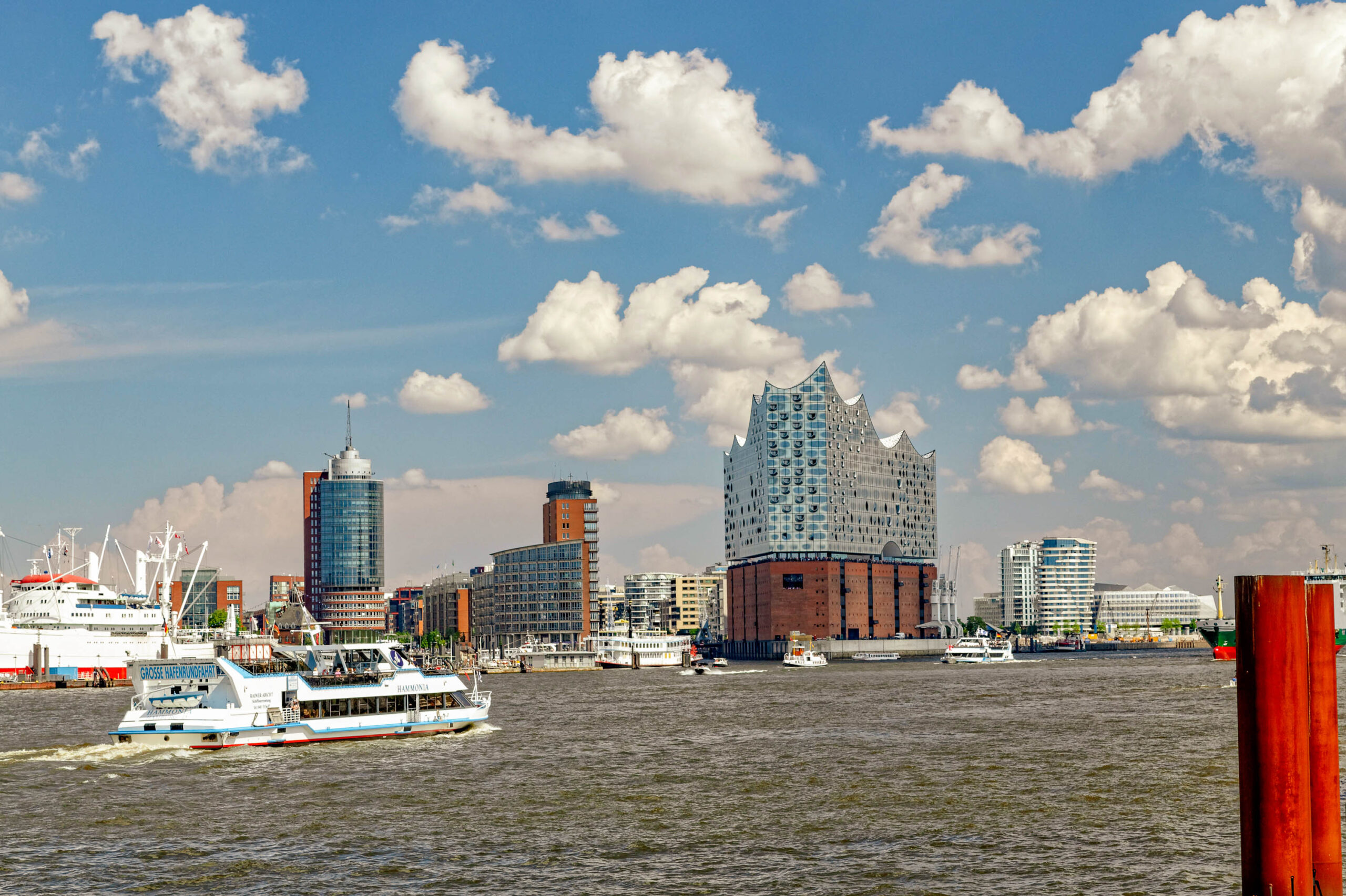 Die Aufnahme zeigt eine malerische Ansicht der Hamburger Skyline vom Hafen aus, aufgenommen im Mai 2018. Ein Passagierschiff, vermutlich auf dem Weg aus dem Hafen, dominiert den Vordergrund. Die Elbphilharmonie, mit ihrer markanten, wellenförmigen Dachkonstruktion, ist ein zentrales Element der Skyline. Verschiedene moderne Hochhäuser und Gebäude ergänzen das Stadtbild. Der Himmel ist strahlend blau und von weißen, flockigen Wolken durchzogen, was der Szene eine sommerliche Atmosphäre verleiht. Das Wasser des Hafens spiegelt das Licht und die Gebäude wider, was die Komposition zusätzlich belebt.