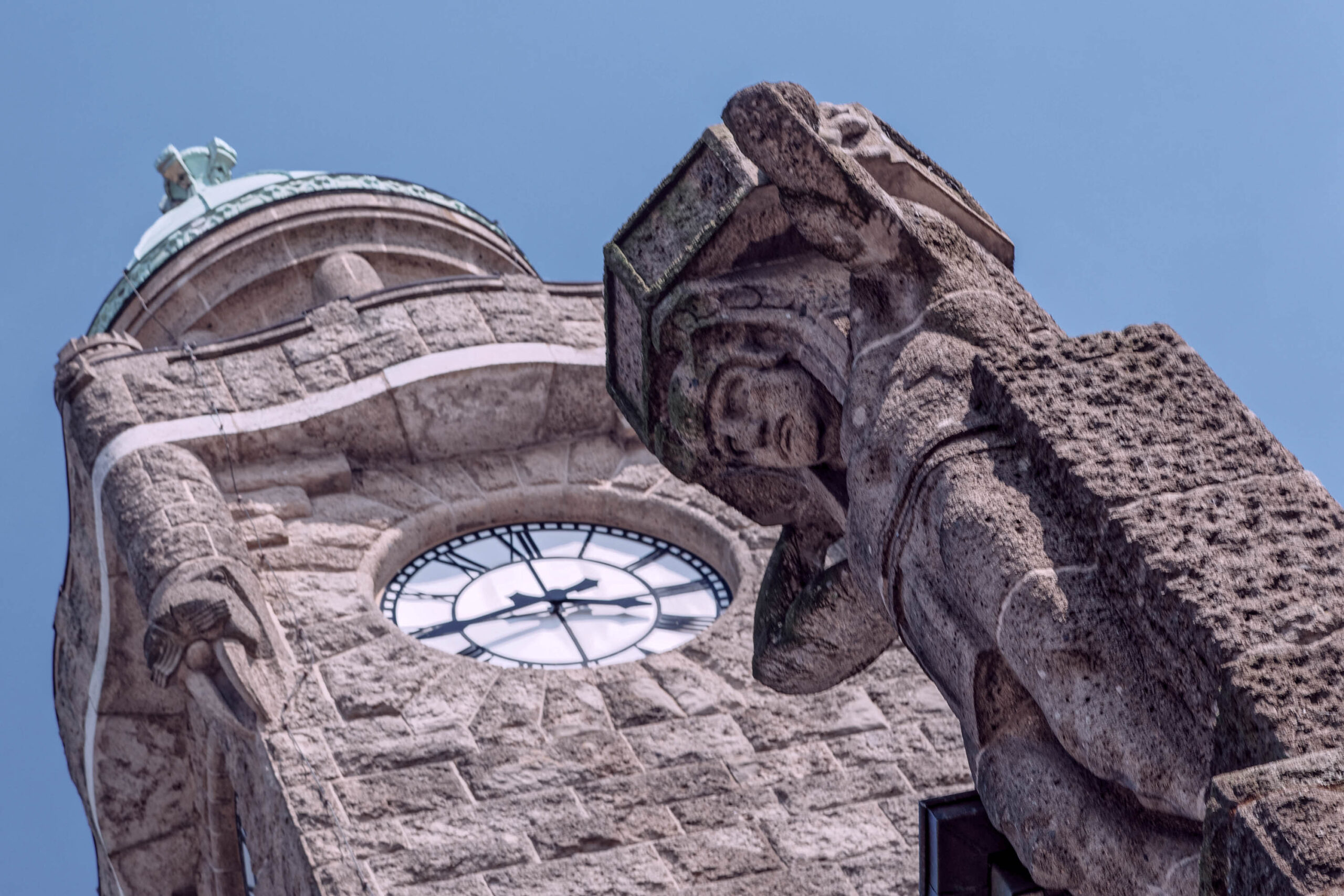 Die Aufnahme zeigt einen Ausschnitt des Turms des Hamburger Hauptbahnhofs. Der Turm ist aus hellem Sandstein gebaut und zeigt eine detaillierte Skulptur eines Drachen oder eines ähnlichen Fabelwesens. Im Hintergrund ist eine Uhr mit römischen Ziffern zu sehen. Der Himmel ist blau und wolkenlos.