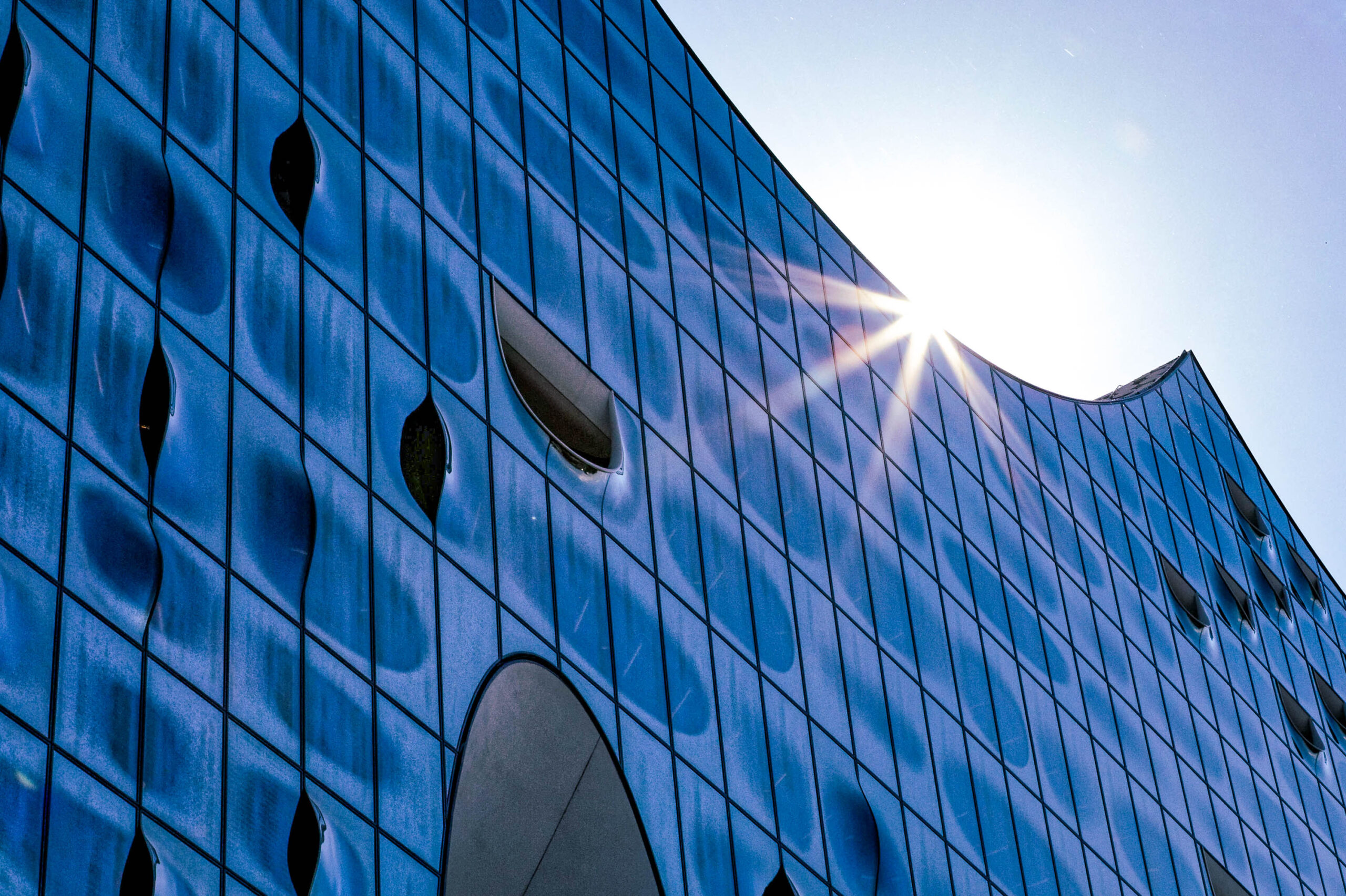 Die Aufnahme zeigt eine Detailaufnahme der Fassade der Elbphilharmonie in Hamburg. Die Fassade besteht aus einer Wellenstruktur aus Glasplatten, die das Sonnenlicht stark reflektieren. Die Glasplatten sind in einem tiefen Blauton gehalten, der durch die Reflexionen und die Beleuchtung verstärkt wird. Die Fassade ist durchbrochen von runden und ovalen Öffnungen, die Fenster darstellen. Die Aufnahme ist aus einem niedrigen Winkel aufgenommen, wodurch die Größe und die Dynamik der Fassade betont werden. Der Himmel ist blau und wolkenlos, was die Farben der Fassade hervorhebt. Die Aufnahme ist scharf und detailliert, wodurch die Textur und die Komplexität der Fassade gut zur Geltung kommen.