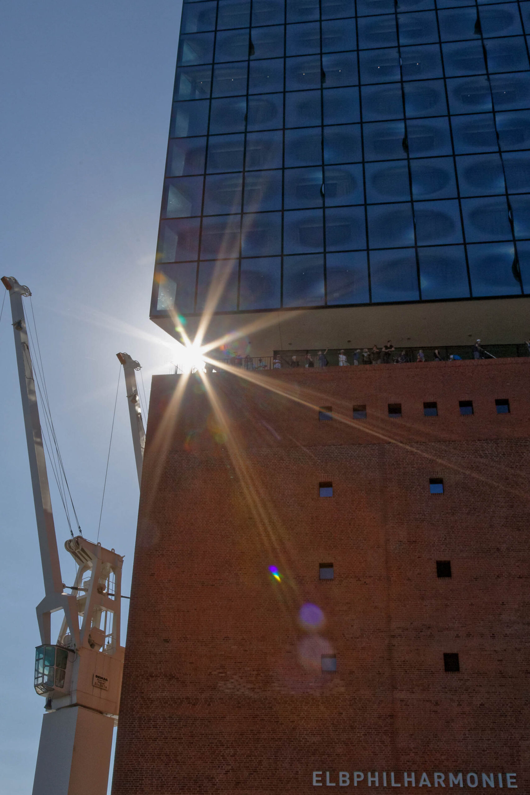Das Foto zeigt eine ungewöhnliche Perspektive auf die Elbphilharmonie in Hamburg. Ein großer Mobilkran dominiert den linken Bildrand und steht vor der Fassade des Konzerthauses. Die Fassade besteht aus unzähligen rechteckigen Fenstern, die das Sonnenlicht reflektieren und einen fast blendenden Effekt erzeugen. Die Sonne scheint direkt auf das Gebäude und erzeugt lange Lichtstrahlen, die sich über das Bild erstrecken. Der Himmel ist klar und blau, was die Szene noch heller und kontrastreicher macht. Der Mai 2018 deutet darauf hin, dass das Foto während der Bauphase der Elbphilharmonie aufgenommen wurde, als noch Bauarbeiten stattfanden.