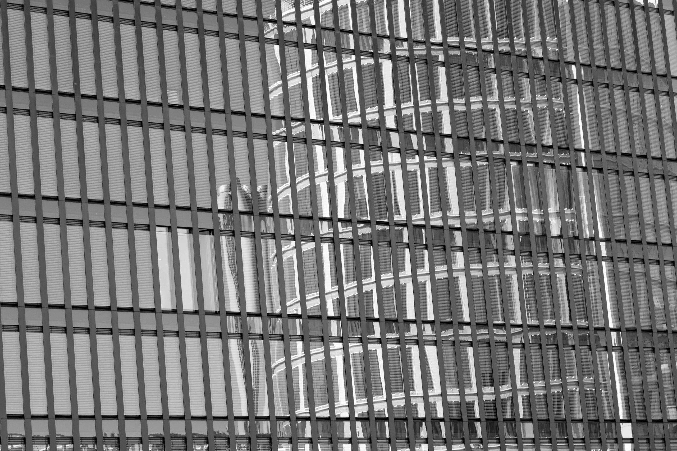 Das vorliegende Schwarz-Weiß-Foto zeigt eine faszinierende Abstraktion der Elbphilharmonie in Hamburg. Die Reflexion des Konzerthauses ist in die Glasfassade eines modernen Gebäudes eingebettet. Die vertikalen Streifen der Fassade brechen das Bild auf und erzeugen ein fragmentiertes, fast kaleidoskopisches Muster. Die Komposition ist dynamisch und spielt mit Licht und Schatten, wodurch eine interessante visuelle Spannung entsteht. Der Kontrast zwischen den klaren Linien der Fassade und der organischen Form der Elbphilharmonie ist bemerkenswert. Das Foto fängt die moderne Architektur Hamburgs auf eine künstlerische und ungewöhnliche Weise ein.