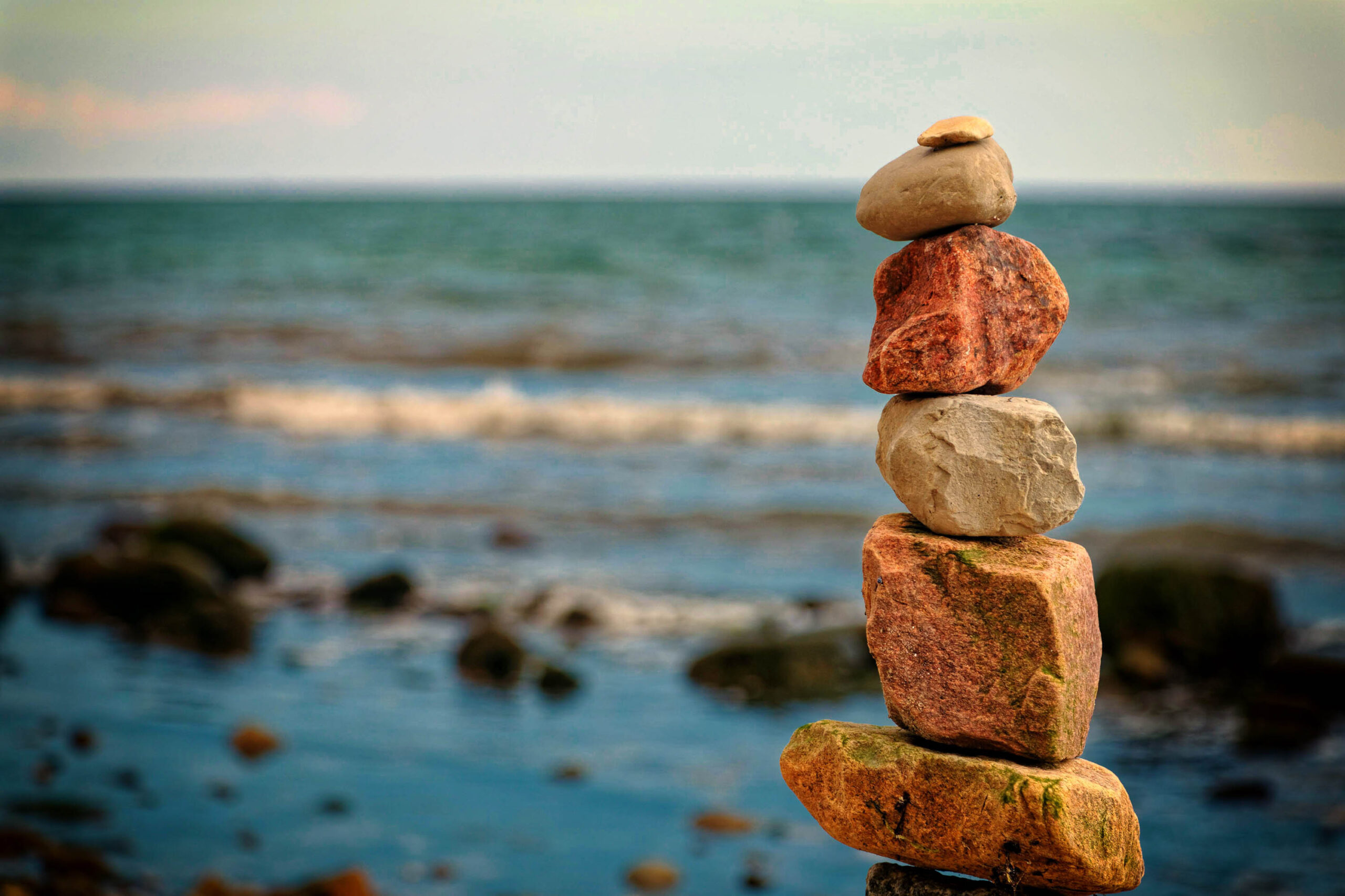 Das Bild zeigt einen sorgfältig gestapelten Turm aus Felsen. Die Felsen variieren in Farbe und Größe, von rötlich-braun bis zu hellgrau. Der Turm steht auf einem sandigen Untergrund, der von kleinen Steinen und Muscheln übersät ist. Im Hintergrund erstreckt sich das Meer, dessen Oberfläche in sanften Wellen schimmert. Der Himmel ist leicht bewölkt und trägt zu einer ruhigen Atmosphäre bei. Die Schärfentiefe ist gering, was den Fokus auf den Felsensturm lenkt und den Hintergrund verschwimmt.