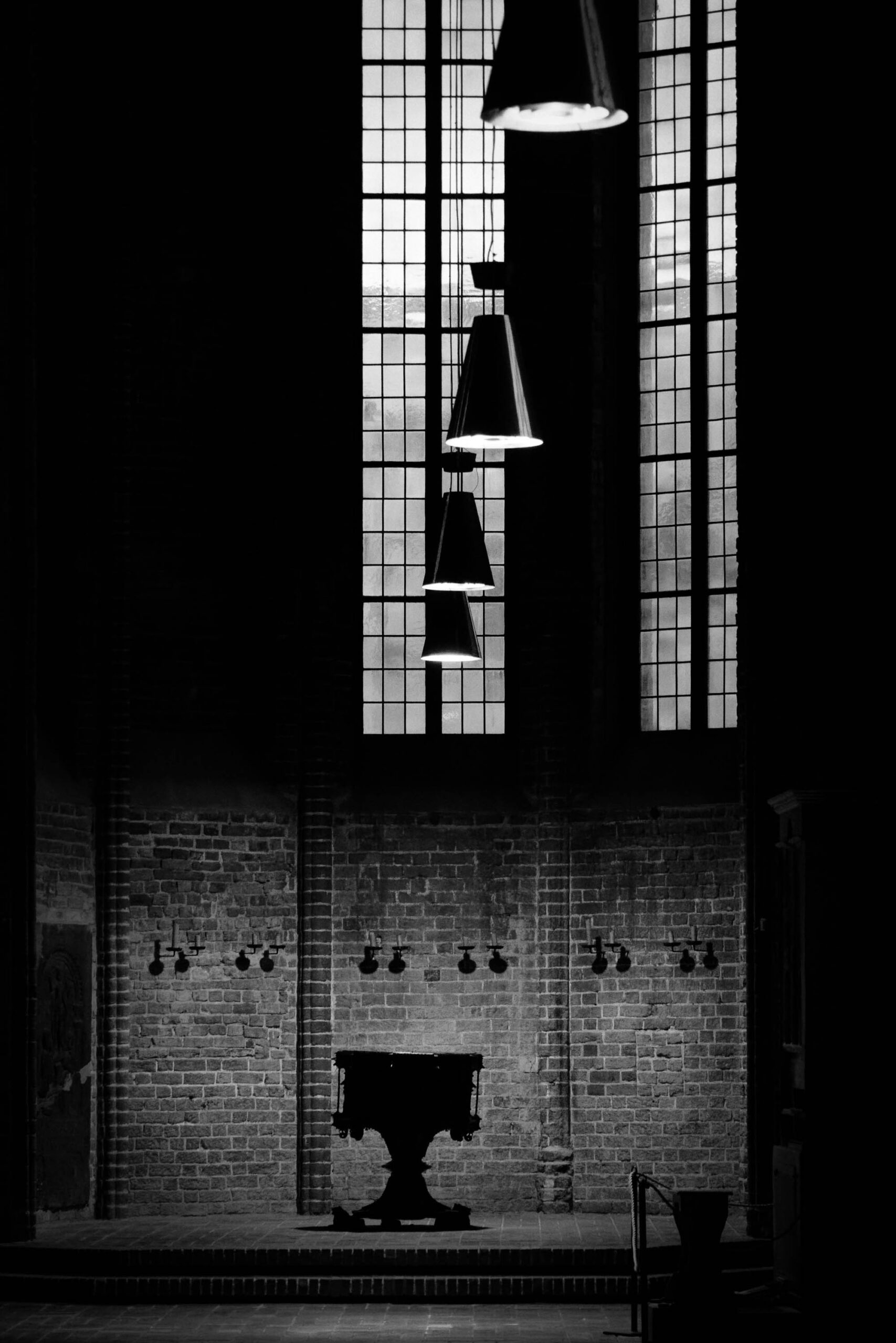 Das Foto zeigt eine düstere, kontrastreiche Innenansicht einer Kirche. Das Licht fällt durch hohe, schmale Fenster und beleuchtet einen Altar. Die Architektur ist von Backsteinmauern und einem schlichten, modernen Design geprägt. Die Atmosphäre ist ruhig und kontemplativ.