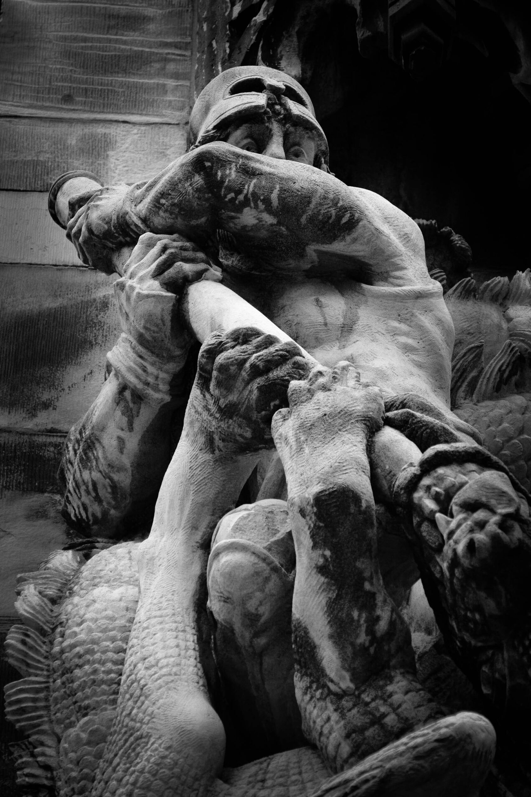 Die Aufnahme zeigt eine detaillierte Schwarz-Weiß-Fotografie einer Skulptur, vermutlich aus Stein oder Marmor. Die Skulptur stellt einen Mann in einer dramatischen, dynamischen Pose dar. Er scheint zu fallen oder sich zu winden, wobei sein Gesichtsausdruck Anstrengung oder Schmerz andeutet. Die Skulptur ist in einem klassischen Stil gehalten und zeigt eine hohe Detailgenauigkeit in der Darstellung der Anatomie und der Gewänderfalten. Der Hintergrund ist unscharf und besteht aus einer Wand oder einer Mauer. Die Beleuchtung ist dramatisch, mit starken Hell-Dunkel-Kontrasten, die die Dynamik der Pose und die Details der Skulptur hervorheben. Die Aufnahme ist vermutlich in einem Park oder einem öffentlichen Raum aufgenommen worden, da sich im Hintergrund eine Mauer oder eine Wand zu erkennen ist. Die Komposition ist auf die Skulptur fokussiert, die den Hauptteil des Bildes ausmacht. Die Aufnahme ist vermutlich in einem Park oder einem öffentlichen Raum aufgenommen worden, da sich im Hintergrund eine Mauer oder eine Wand zu erkennen ist.