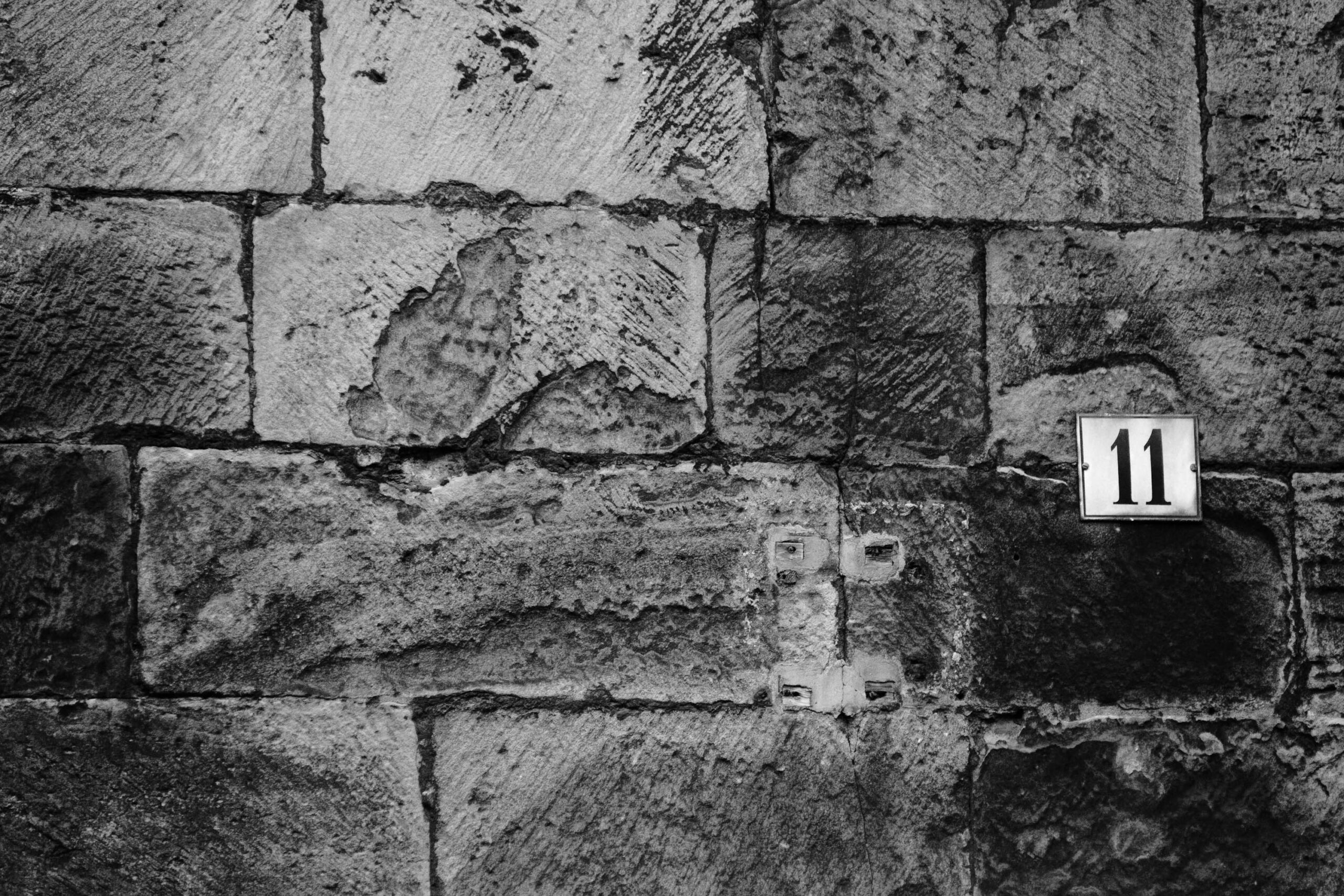 Das Foto zeigt eine Mauer aus grob behauenen Steinblöcken. Die Mauer ist in verschiedenen Grautönen dargestellt, was die Textur und die Unebenheiten der Steine hervorhebt. Eine Hausnummer '11' ist in einem rechteckigen Schild angebracht, das sich deutlich von der rauen Oberfläche der Mauer abhebt. Das Licht scheint von oben zu kommen und wirft Schatten, die die Tiefe der Mauerstruktur betonen. Die Komposition ist einfach und konzentriert sich auf die Textur und die geometrische Form der Mauer und des Schildes.