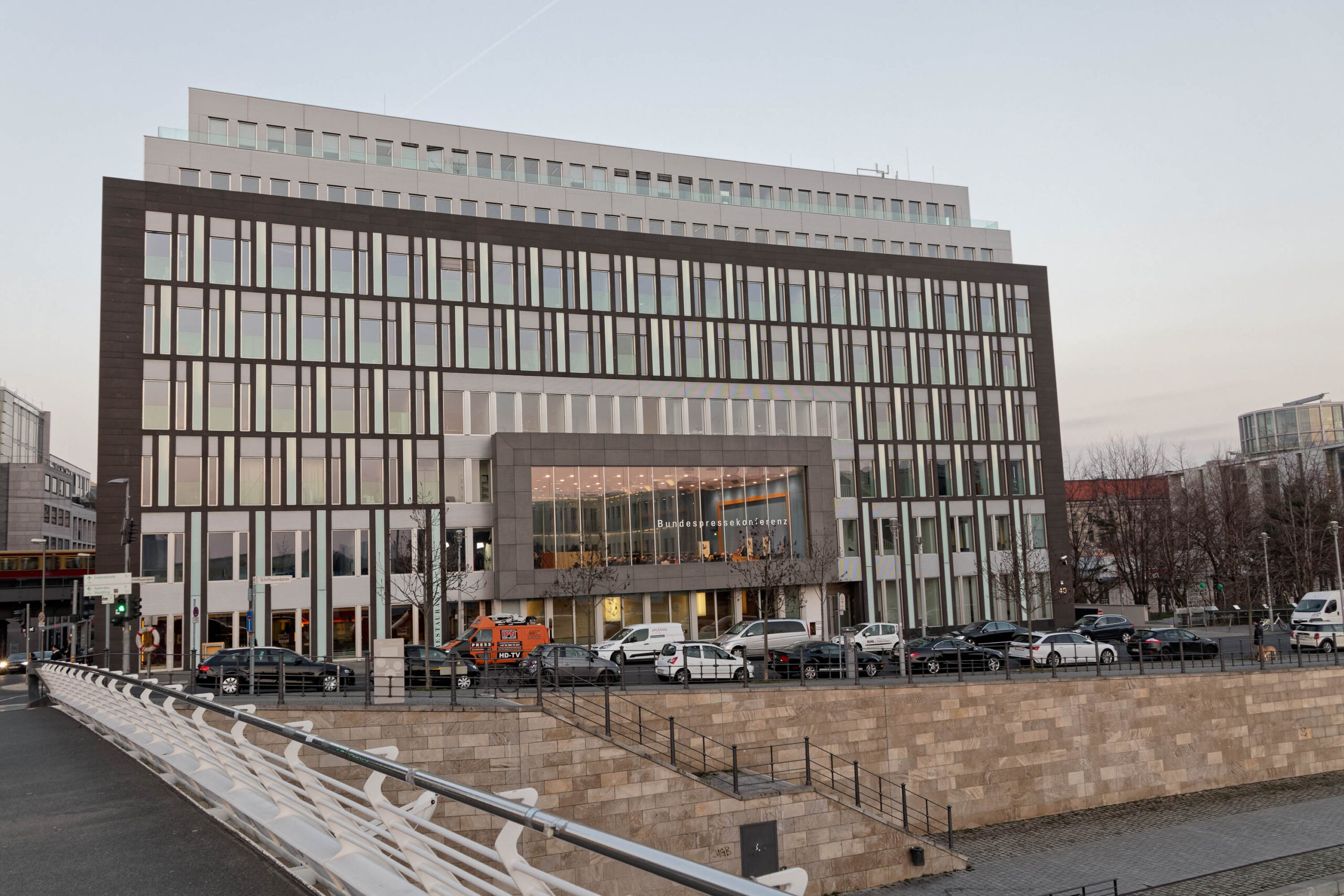 Das Foto zeigt eine Frontansicht des modernen Gebäudes der Bundespressekonferenz in Berlin. Das Gebäude ist ein rechteckiger, mehrstöckiger Bau mit einer Fassade aus dunklen horizontalen Lamellen und großen Fensterflächen. Die Fassade wirkt durch die Anordnung der Elemente sehr strukturiert und geometrisch. Vor dem Gebäude befindet sich eine breite Promenade, die von einer Steinmauer und einem Geländer abgetrennt ist. Auf der Promenade parken mehrere Fahrzeuge, darunter PKWs und ein Lieferwagen. Im Hintergrund sind weitere Gebäude und Bäume zu sehen. Der Himmel ist leicht bewölkt und die Lichtverhältnisse deuten auf den späten Nachmittag hin.