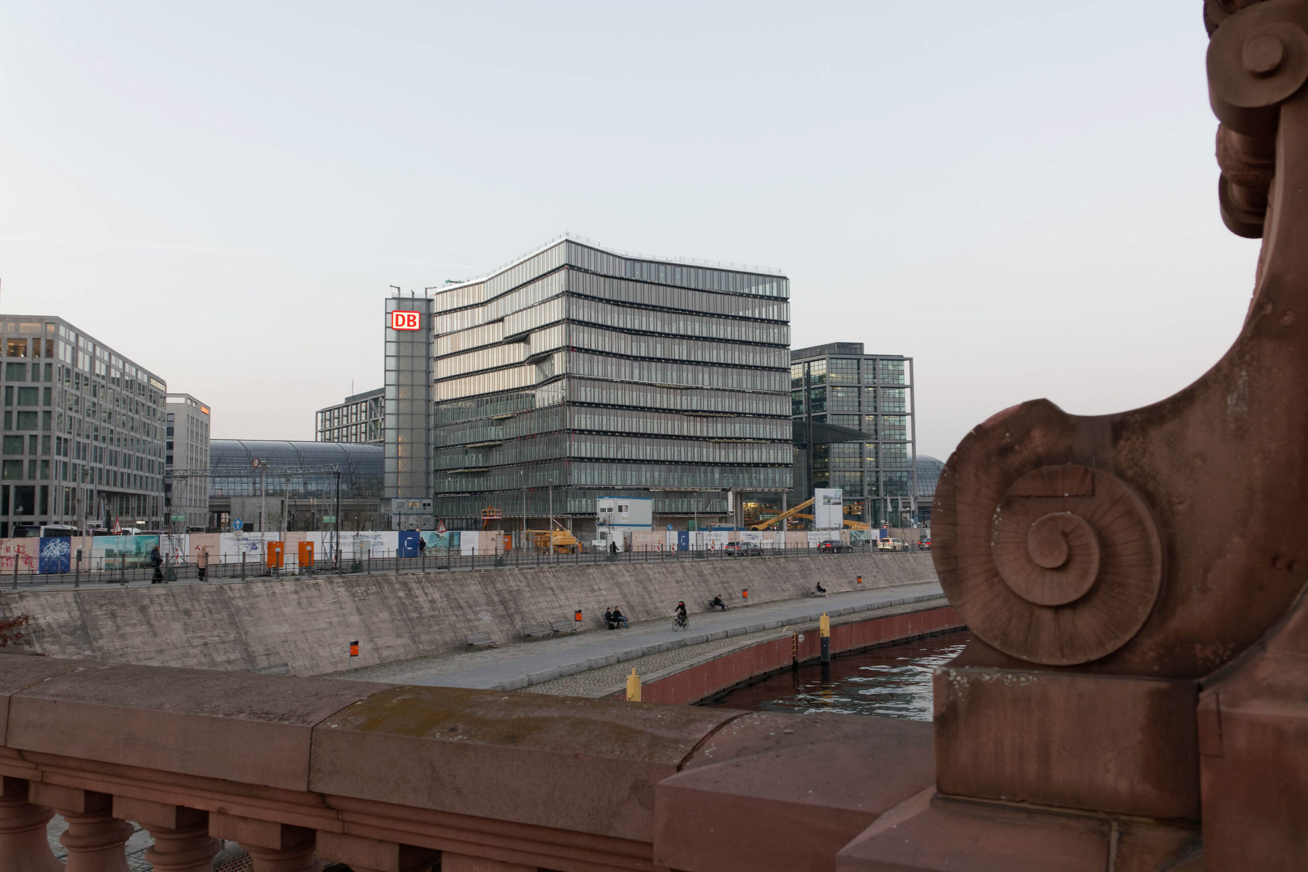 Die Aufnahme zeigt eine moderne Skyline in Berlin, dominiert vom markanten Bürogebäude der Deutschen Bahn (DB). Im Vordergrund fließt ein Fluss, der von einem Ufer mit einem schmalen Weg gesäumt ist. Ein alter Steinbalustrade rahmen die Szene und verleiht ihr eine historische Note. Der Himmel ist bedeckt, was eine gedämpfte Stimmung erzeugt. Die Architektur der Gebäude ist modern und minimalistisch, mit großen Glasflächen, die das Licht reflektieren. Die Szene vermittelt einen Eindruck von urbaner Entwicklung und historischer Kontraste.