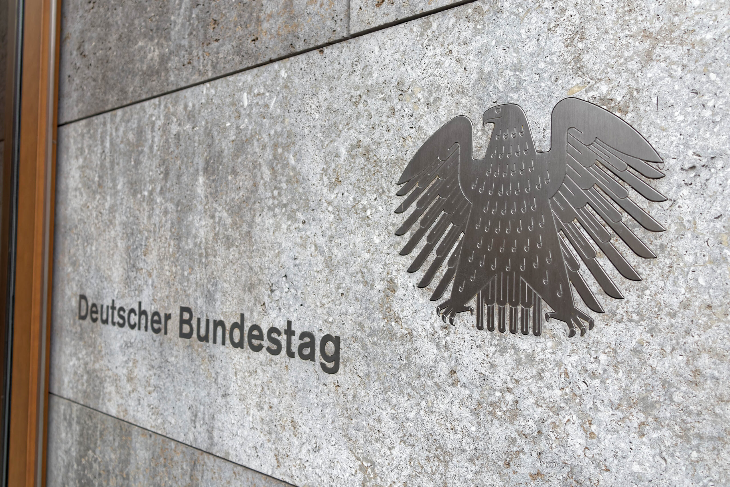 Das Bild zeigt eine Detailaufnahme der Fassade des Deutschen Bundestages in Berlin. Im Vordergrund befindet sich ein stilisiertes Abbild des Bundesadlers, das aus Metall gefertigt ist und auf einer grauen Betonwand angebracht ist. Darunter befindet sich der Schriftzug 'Deutscher Bundestag'. Die Aufnahme ist aus einer Perspektive aufgenommen, die den Adler und den Schriftzug in den Fokus rückt. Die Textur der Betonwand ist deutlich erkennbar.