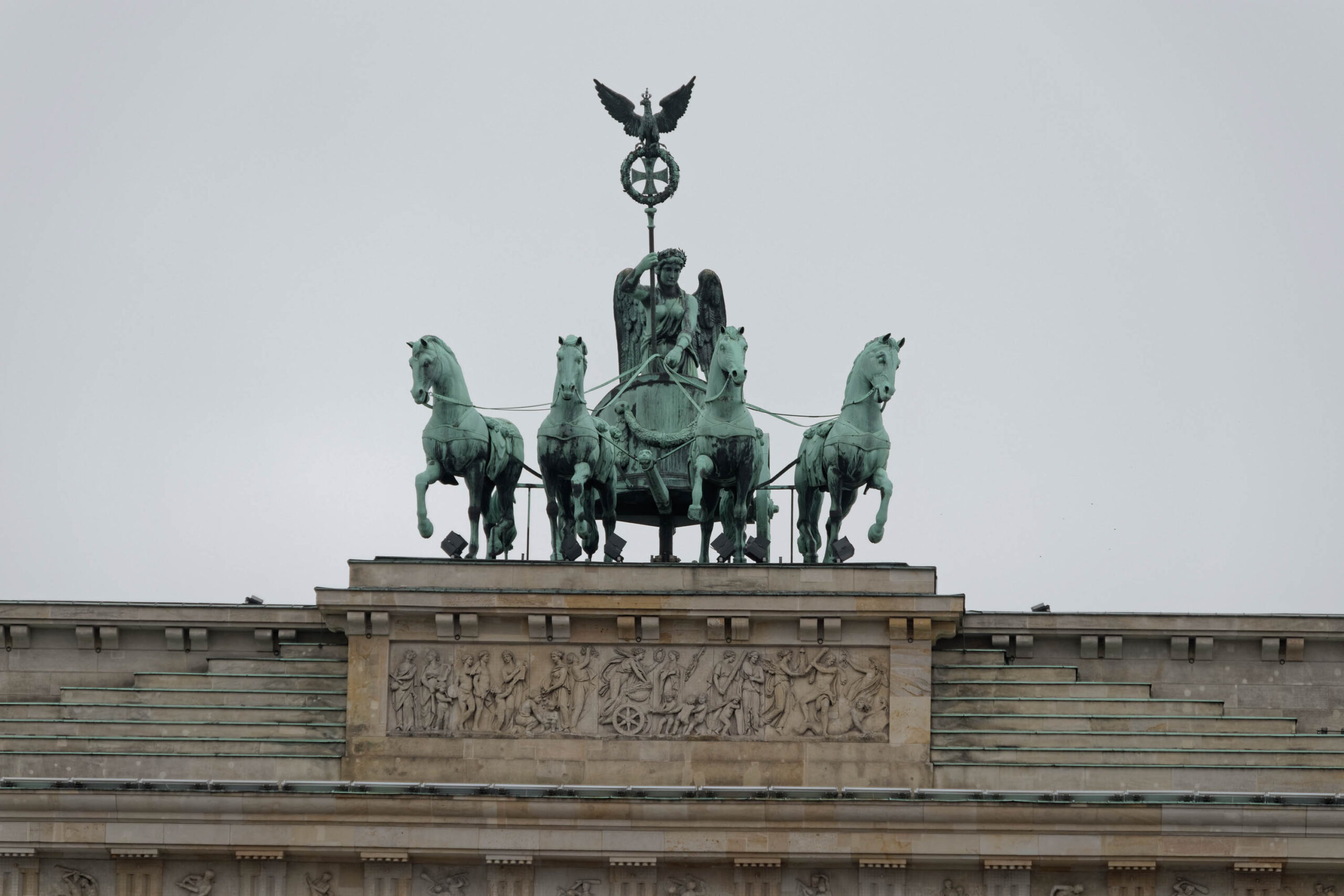Das Foto zeigt die Quadriga, die auf dem Brandenburger Tor in Berlin thront. Die Quadriga ist eine Skulptur, die eine viergespannte Kutsche mit einer Siegesgöttin (Victoria) darstellt. Die Skulptur ist aus Bronze und hat eine grüne Patina. Der Himmel ist bewölkt und das Licht ist diffus.