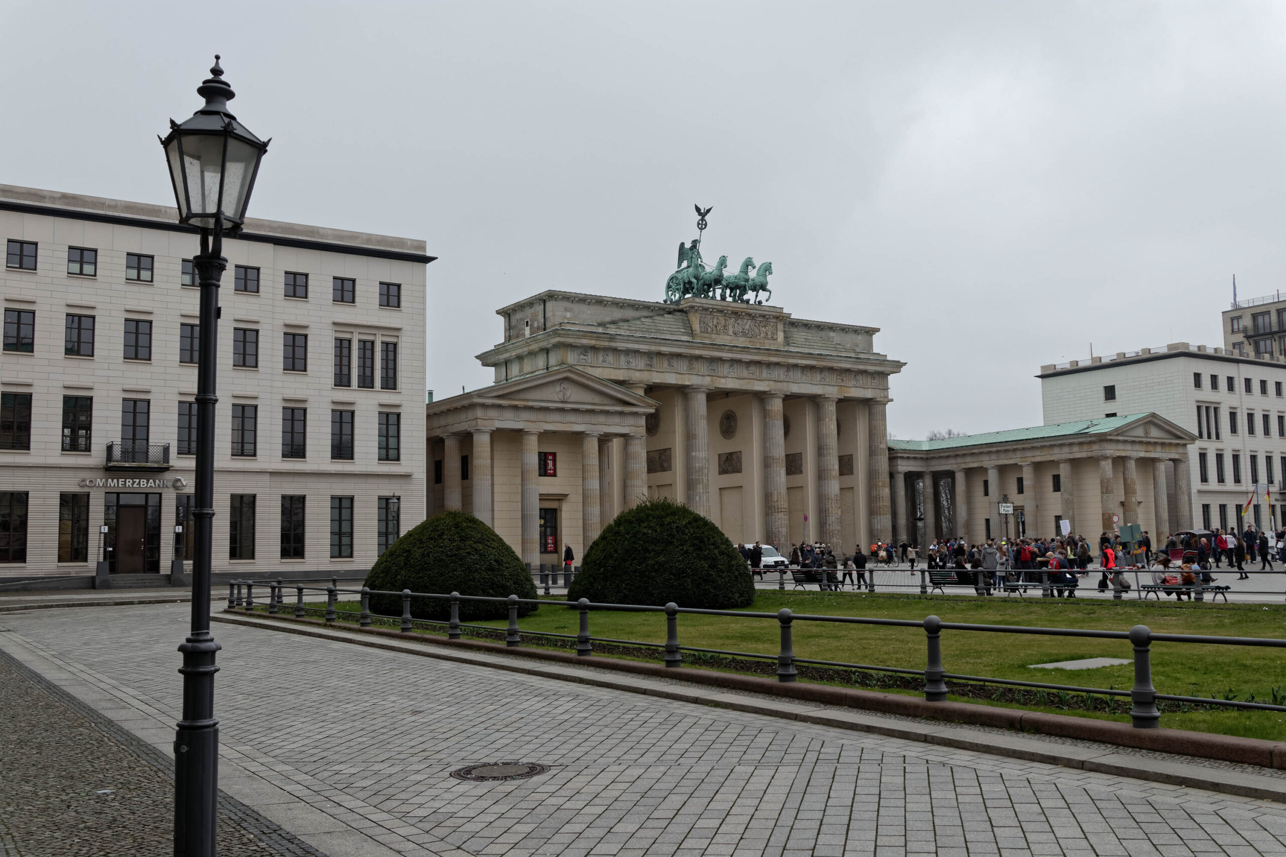 Das Foto zeigt eine Szene in Berlin im März 2019. Im Vordergrund befindet sich ein gepflasterter Platz mit einem historischen Laternenpfahl. Im Hintergrund dominiert das Brandenburger Tor, dessen Quadriga im Regen steht. Links des Brandenburger Tors befindet sich ein klassizistisches Gebäude, vermutlich ein Teil des Museumsblocks. Mehrere Personen sitzen auf Bänken und beobachten die Szene. Der Himmel ist bedeckt und es regnet.