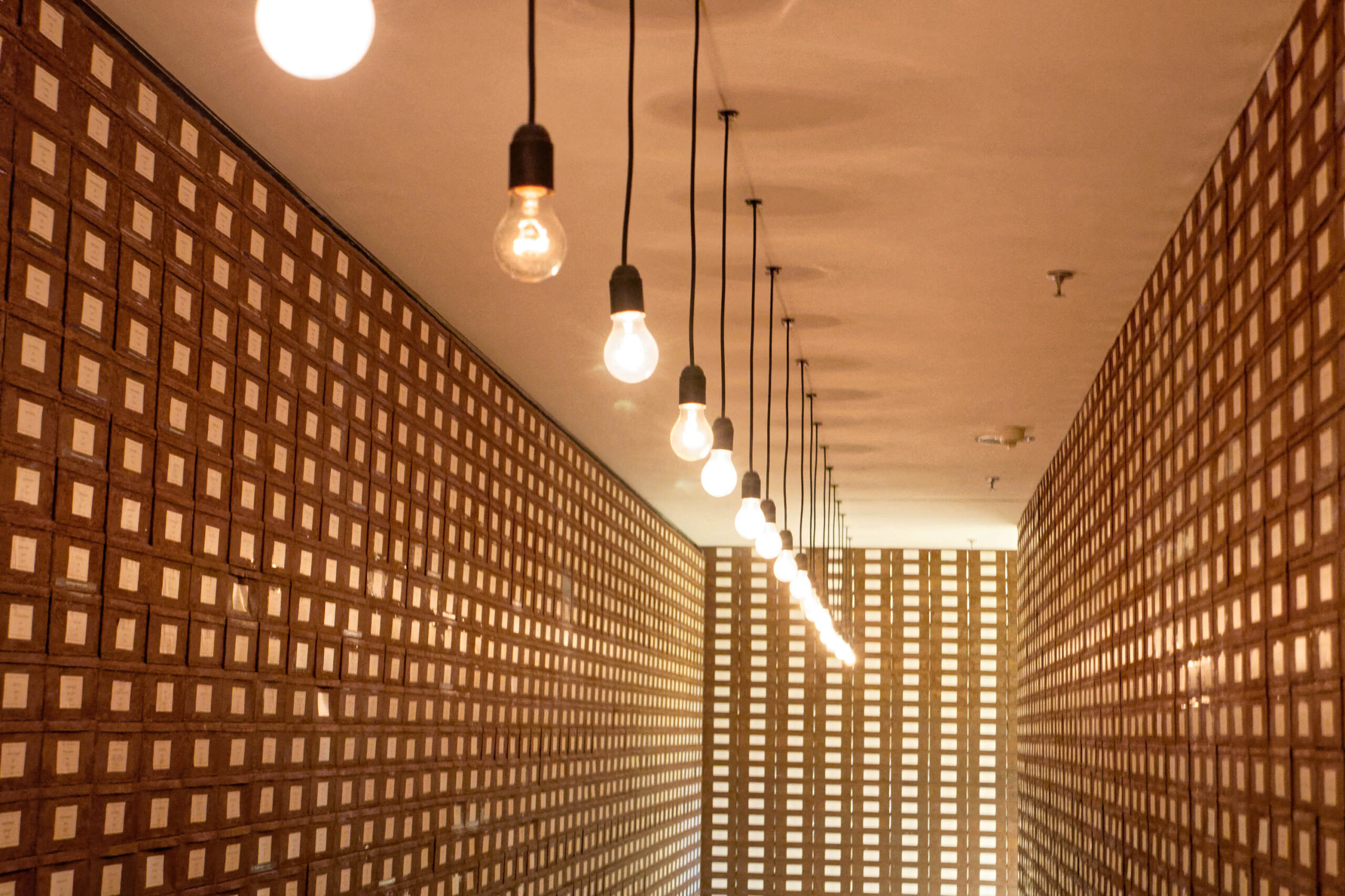 Das Foto zeigt einen langen, schmalen Gang, dessen Wände von unzähligen Aktenordnern gefüllt sind. Die Ordner sind in einem Raster angeordnet und erzeugen ein geometrisches Muster.  Die Beleuchtung besteht aus hängenden Glühbirnen, die einen warmen, goldenen Schein auf die Szene werfen. Die Perspektive betont die Länge und Enge des Ganges. Die Atmosphäre ist ruhig und fast schon meditativ. Es wirkt wie ein Archiv oder ein Lagerraum.