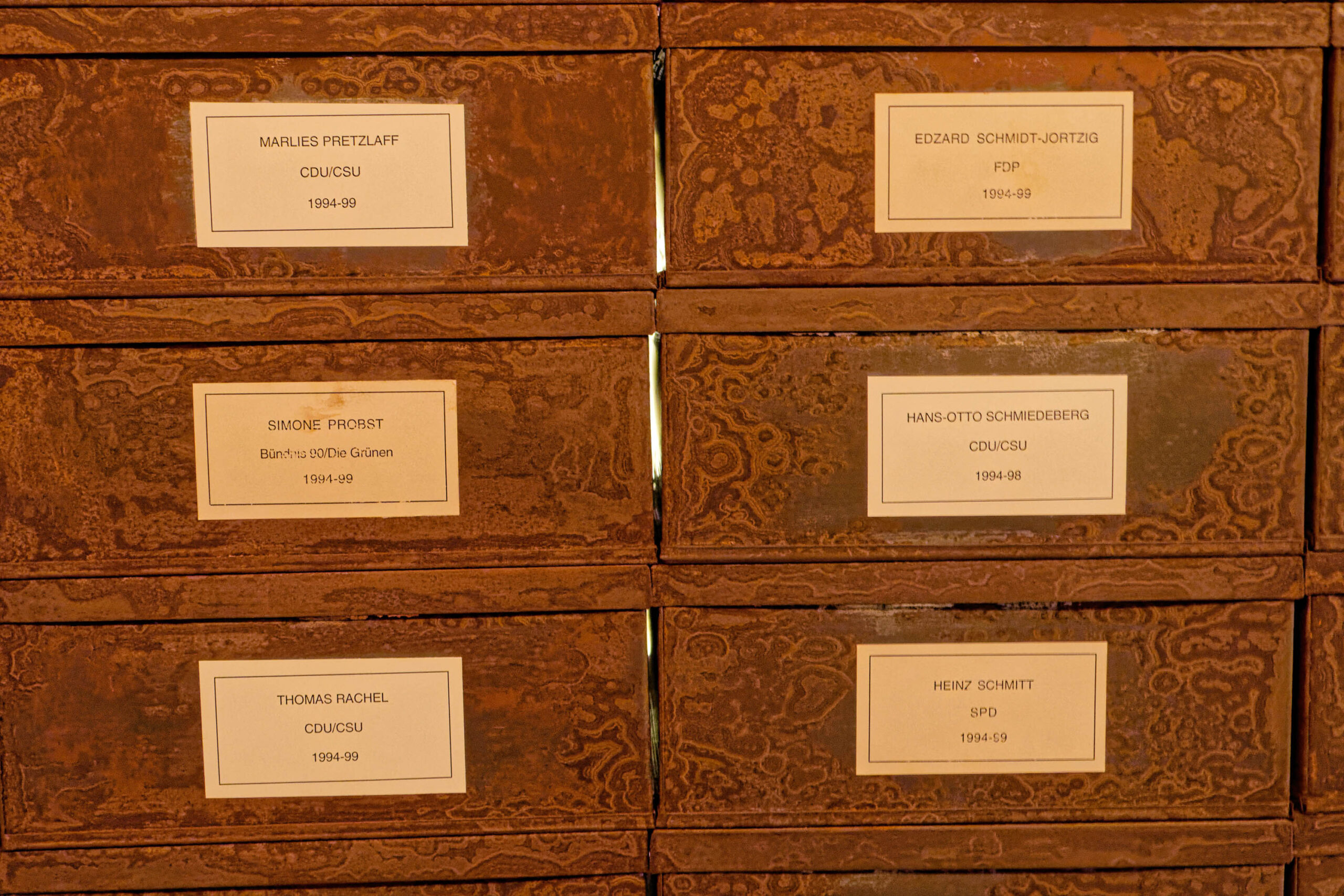 Die Aufnahme zeigt eine Anordnung von Archivkartons, die übereinander gestapelt sind. Die Kartons sind in einem warmen Braunton gehalten und tragen gelbe Etiketten mit Namen und politischen Parteizugehörigkeiten. Die Etiketten sind in einer serifenlosen Schriftart gedruckt und zeigen Namen wie 'Thomas Rachel', 'Simone Probst', 'Marlies Pretzlaff', 'Heinz Schmitt', 'Hans-Otto Schmiedeburg', 'Edzard Schmidt-Jortzig'. Die Namen sind mit den Parteien CDU/CSU, Bündnis 90/Die Grünen, SPD und FDP verbunden. Die Jahreszahlen '1994-99', '1994-98' und '1994-95' sind ebenfalls auf den Etiketten angegeben. Die Anordnung der Kartons deutet auf ein Archiv hin, möglicherweise in einem politischen Kontext.