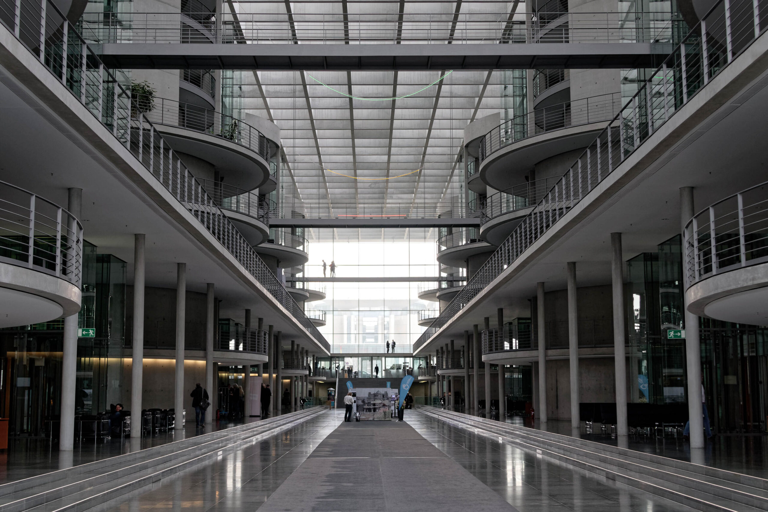 Die Aufnahme zeigt eine beeindruckende Innenansicht des Sony Center in Berlin. Die lange, symmetrische Passage erstreckt sich in die Tiefe und wird von den charakteristischen, kreisförmigen Gebäudestrukturen des Komplexes eingerahmt. Die reflektierende Oberfläche des Bodens verstärkt den Eindruck von Tiefe und Symmetrie. Die Beleuchtung ist diffus und erzeugt eine ruhige, fast meditative Atmosphäre. Mehrere Personen sind in der Ferne zu sehen, die sich in der Passage bewegen.