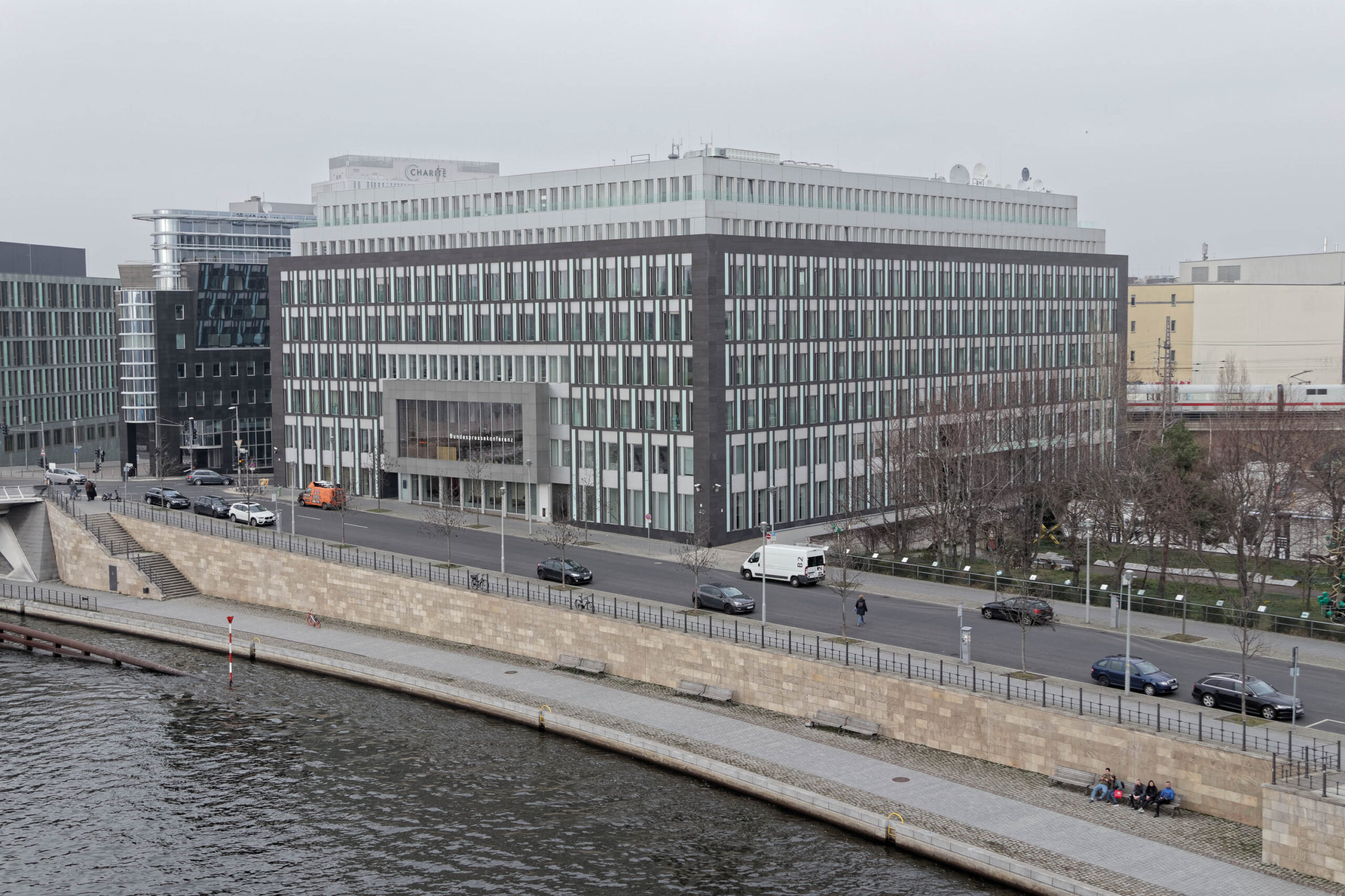 Die Aufnahme zeigt ein modernes, rechteckiges Bürogebäude mit einer Fassade aus dunklen Betonplatten und großen Fensterflächen. Das Gebäude erstreckt sich über mehrere Stockwerke und dominiert die Szene. Im Vordergrund fließt ein Fluss, der von einer Uferbefestigung gesäumt ist. Auf der Straße vor dem Gebäude befinden sich mehrere Fahrzeuge. Der Himmel ist bedeckt und die Lichtverhältnisse sind diffus.