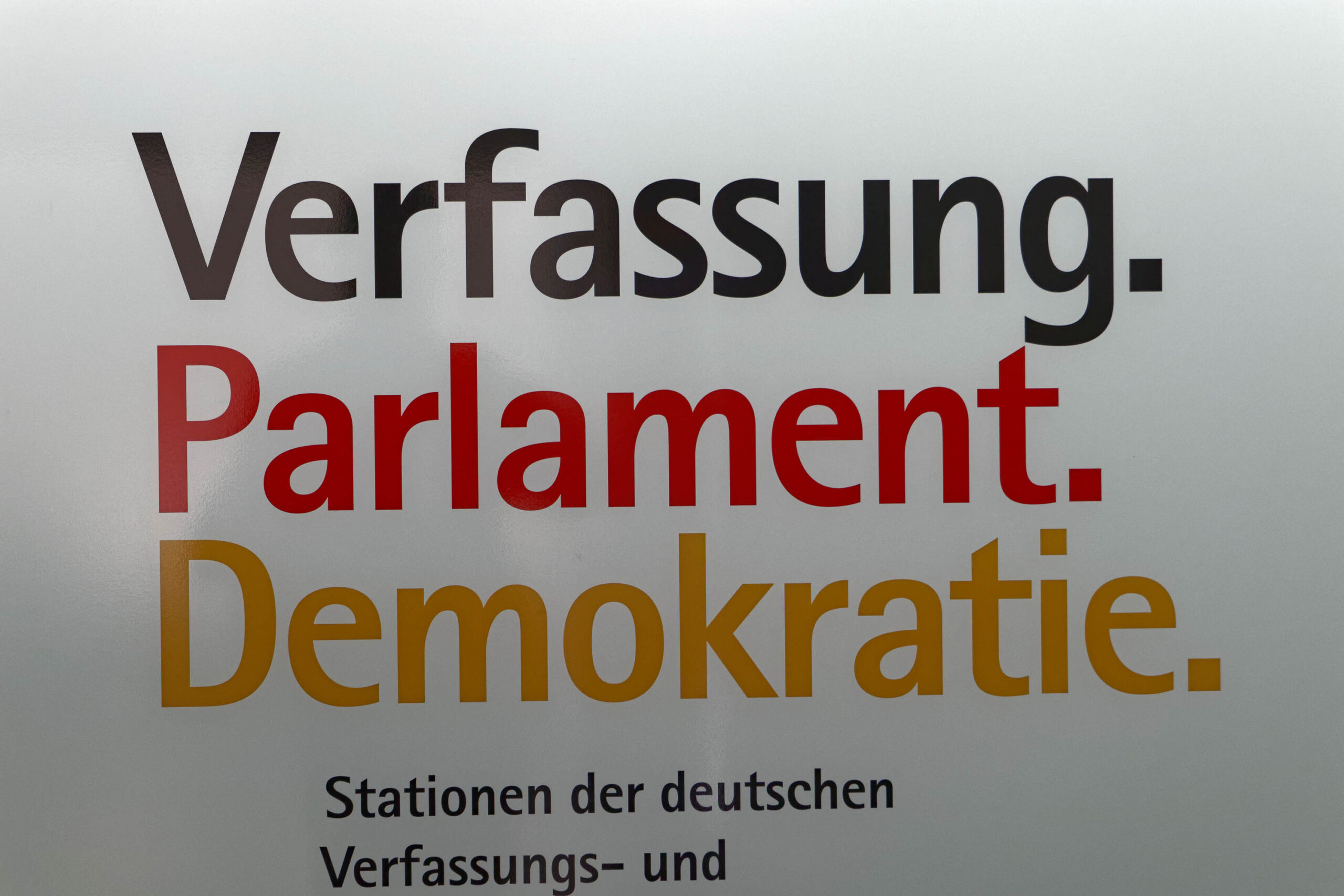 Die Fotografie zeigt eine Nahaufnahme eines Schildes, das die Wörter „Verfassung“, „Parlament“ und „Demokratie“ in unterschiedlichen Schriftarten und Farben präsentiert. Der Hintergrund ist verschwommen und deutet auf eine Glasfläche hin, was darauf hindeutet, dass das Schild möglicherweise an einem öffentlichen Ort angebracht ist. Die Bildkomposition konzentriert sich auf die Botschaft des Schildes und betont die Bedeutung von Verfassung, Parlament und Demokratie. Der März 2019 Kontext deutet auf eine politische oder gesellschaftliche Veranstaltung hin.