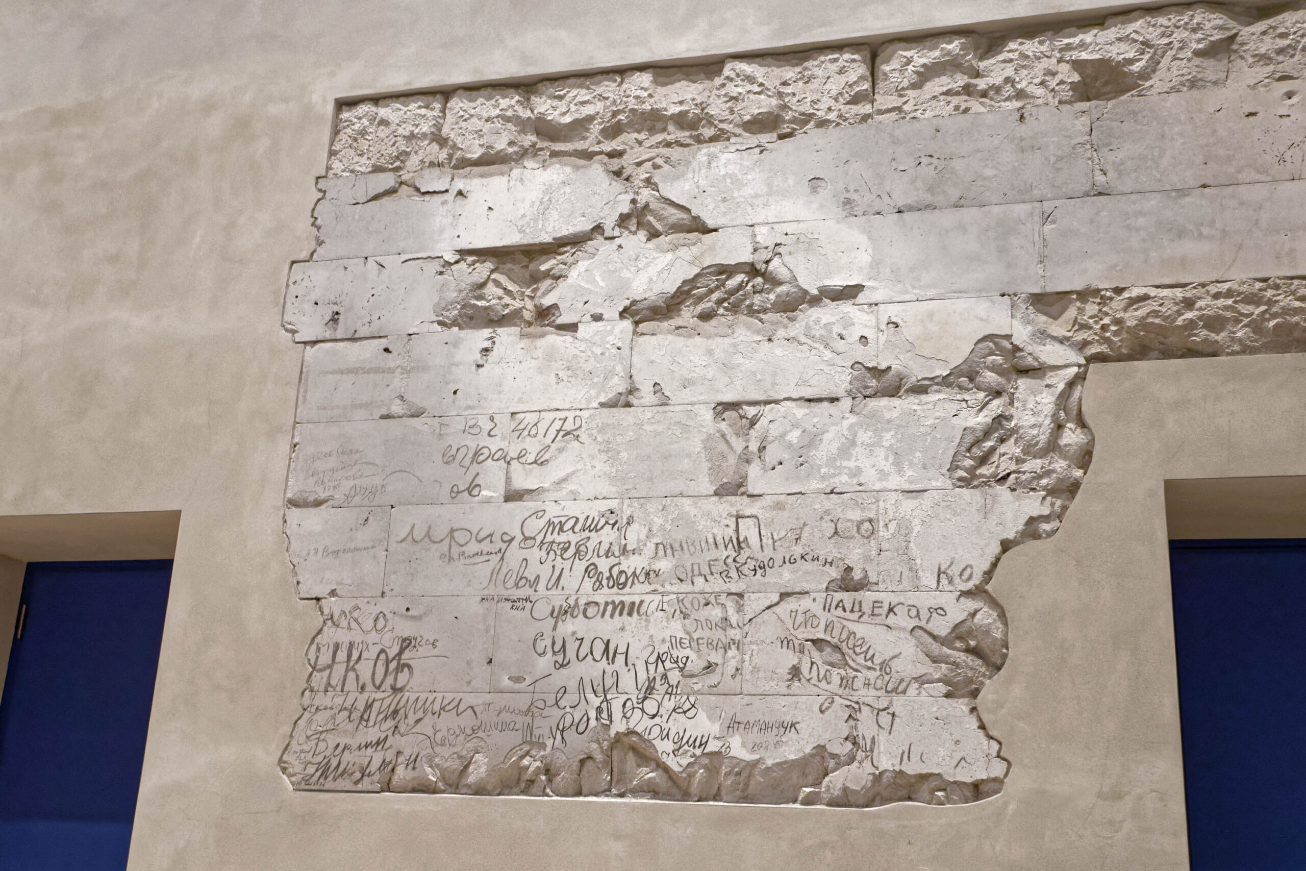 Das Bild zeigt ein erhaltenes Stück der Berliner Mauer, das als Ausstellungsstück an einer weißen Wand angebracht ist. Die Mauer selbst besteht aus Beton und ist mit zahlreichen Graffiti und Unterschriften bedeckt, die in verschiedenen Sprachen, vermutlich Russisch und Deutsch, verfasst wurden. Die Schriftzüge sind teilweise schwer zu entziffern, aber es lassen sich Namen und kurze Botschaften erkennen. Die Mauer ist in einem relativ guten Zustand, aber weist deutliche Abnutzungsspuren auf. Die Beleuchtung betont die Texturen des Betons und die Details der Graffiti. Die Wand, an der die Mauer angebracht ist, wirkt modern und sauber, was einen Kontrast zur rauen Oberfläche der Mauer bildet. Im Hintergrund sind weitere Wände und möglicherweise ein Durchgang zu einem anderen Raum erkennbar.