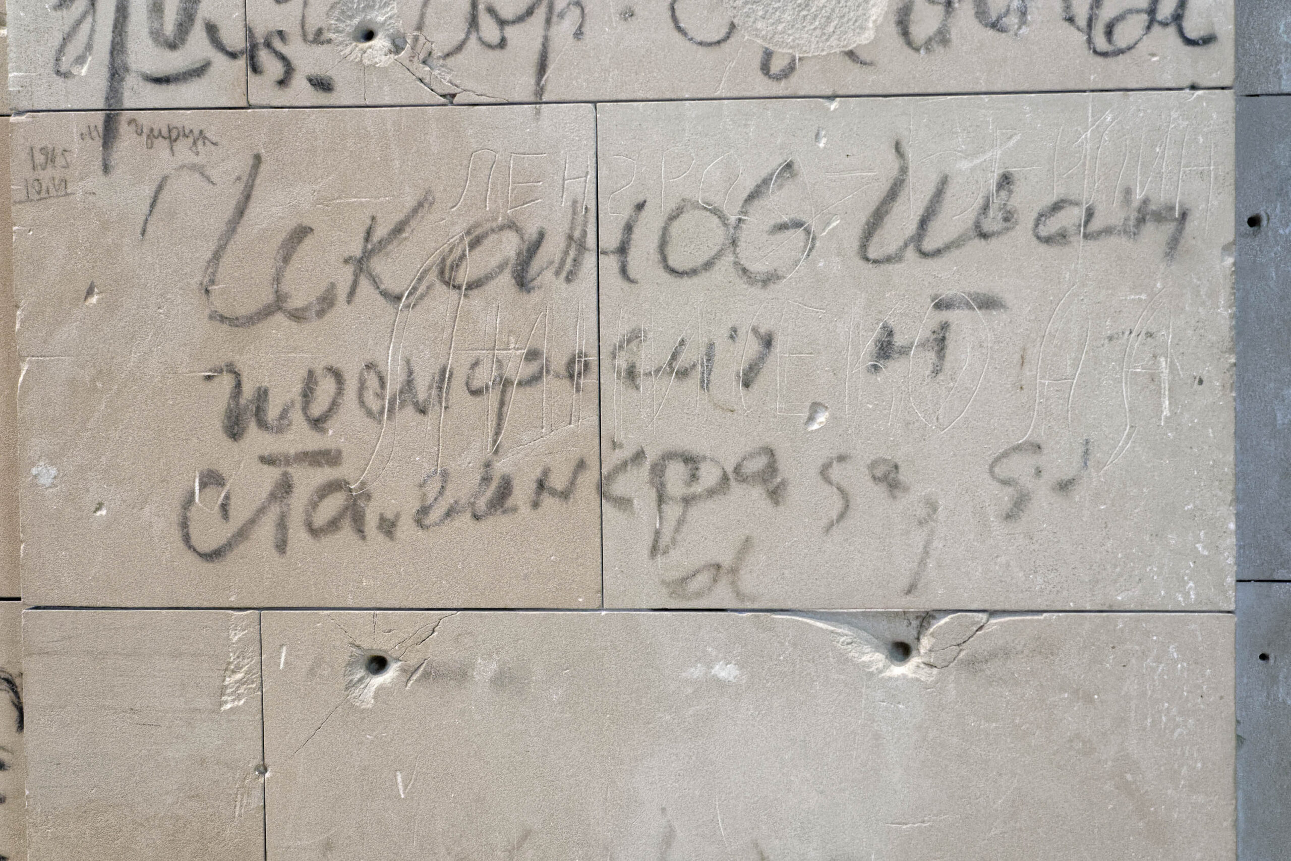 Die Aufnahme zeigt eine Nahaufnahme einer grauen Betonwand. Die Wand ist mit Graffiti in kyrillischer Schrift bedeckt. Es sind mehrere Schusslöcher in der Wand zu sehen. Die Schrift ist in verschiedenen Größen und Stilen geschrieben, was auf unterschiedliche Schreiber und Zeitpunkte hindeutet. Die Graffiti-Texte sind schwer zu entziffern, aber es scheint sich um russische oder belarussische Schrift handelt. Die Wand wirkt rau und abgenutzt, was auf ihre lange Geschichte und die Witterungseinflüsse hindeutet.