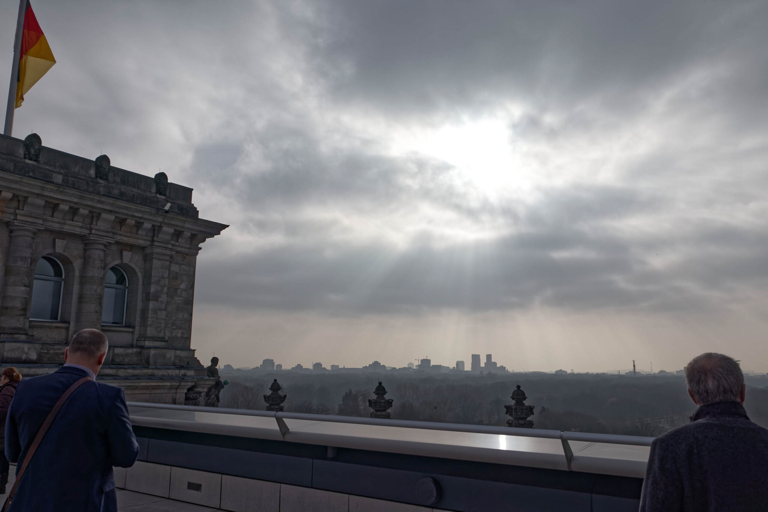 Die Aufnahme zeigt eine Panoramaansicht von Berlin vom Reichstag aus. Zwei Personen stehen am Rand der Kuppel und blicken auf die Stadt. Der Himmel ist bedeckt und lässt das Licht gedämpft wirken. Im Vordergrund sind die Kuppelstrukturen des Reichstags erkennbar, im Hintergrund die Skyline Berlins mit markanten Gebäuden wie dem Fernsehturm. Die Atmosphäre ist ruhig und nachdenklich.