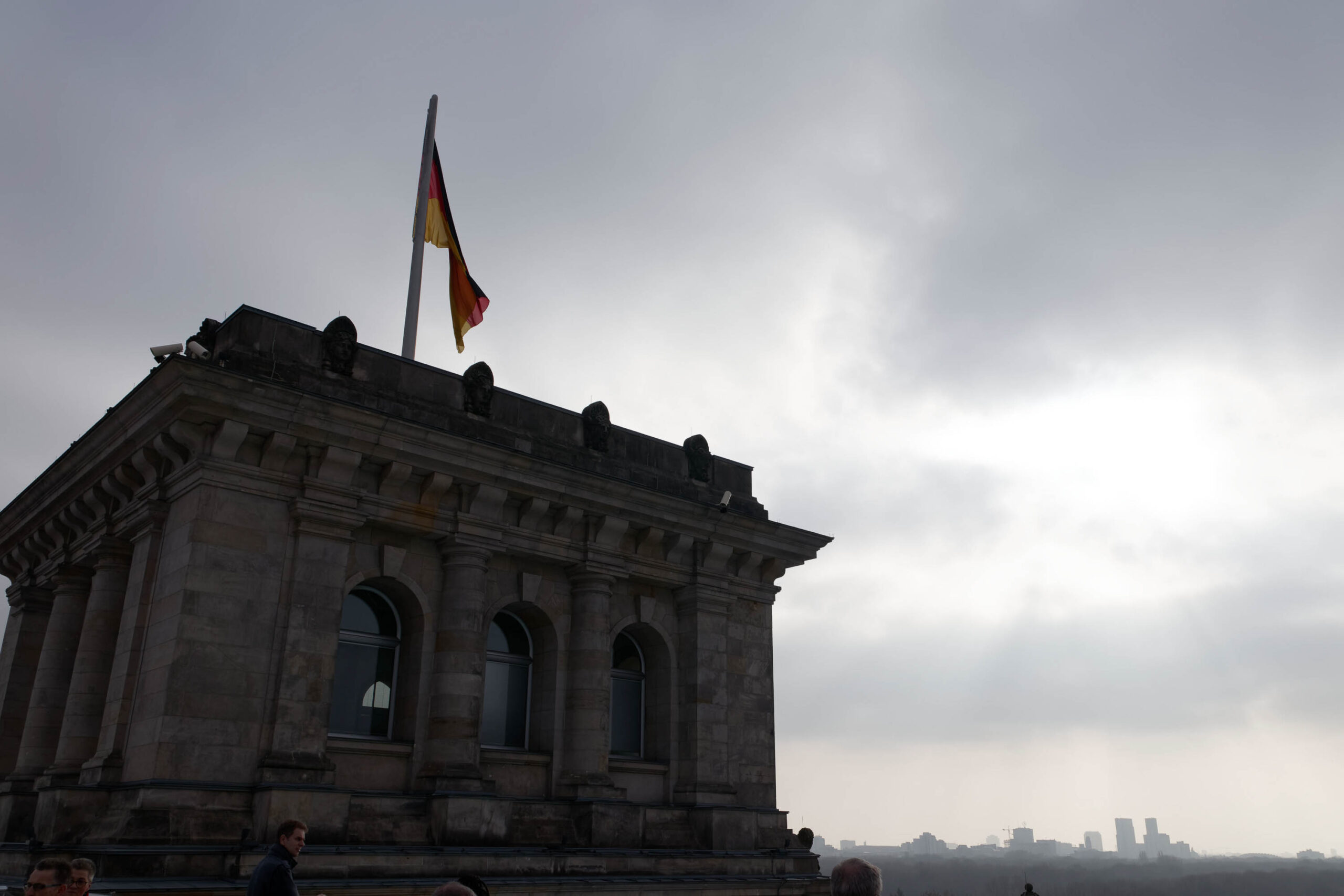 Das Foto zeigt eine partielle Ansicht des Reichstagsgebäudes in Berlin. Der Blickwinkel ist von unten, wodurch die imposante Größe des Gebäudes betont wird. Die deutsche Flagge weht auf dem Flaggenmast. Im Hintergrund ist der Berliner Horizont mit einigen Wolkenkratzern erkennbar. Der Himmel ist bedeckt und die Lichtverhältnisse sind diffus.