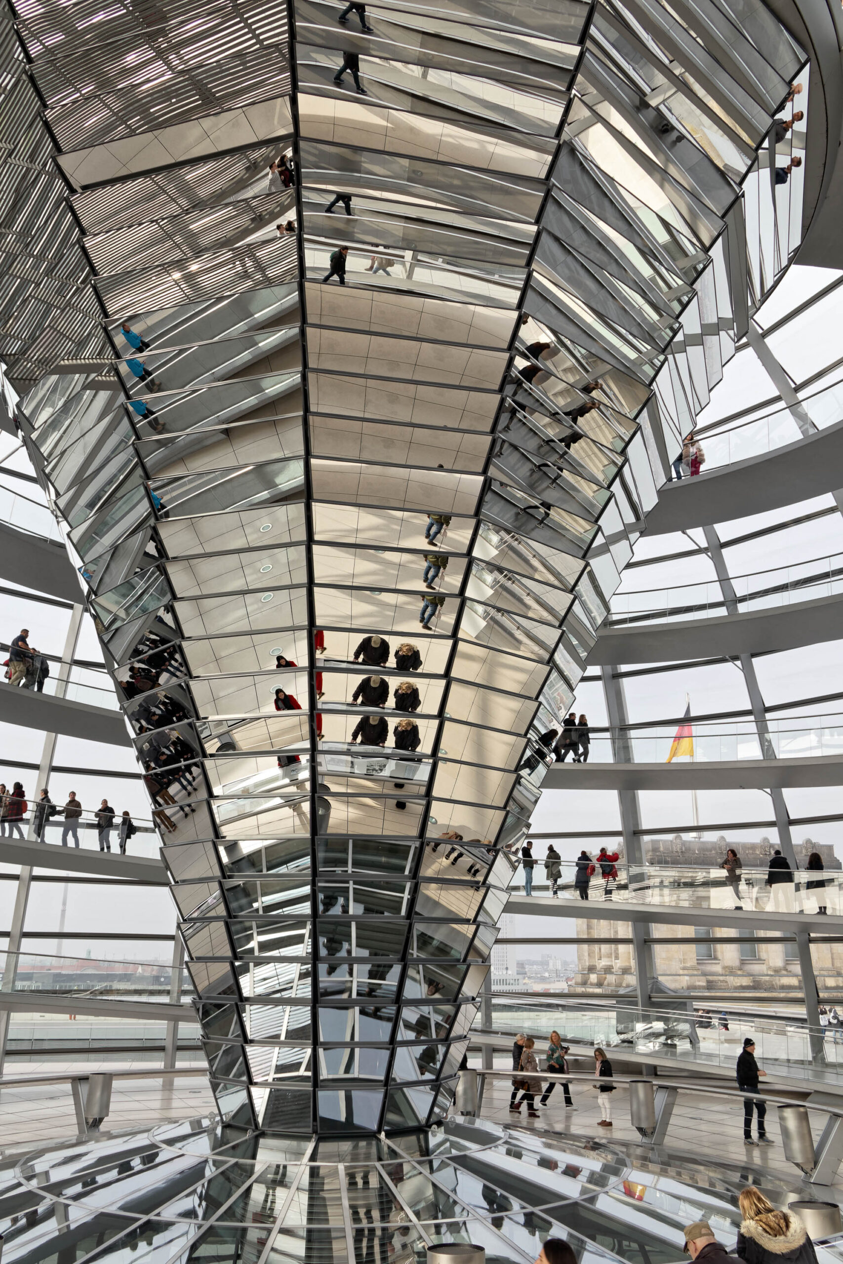 Die Aufnahme zeigt eine Innenansicht der Kuppel des Reichstagsgebäudes in Berlin. Die Kuppel ist ein beeindruckendes Beispiel moderner Architektur, mit ihrer komplexen Stahlkonstruktion und der transparenten Kuppelhaut, die das Sonnenlicht hereinlässt. Mehrere Personen sind in der Kuppel zu sehen, die die Aussicht genießen. Die Perspektive ist von unten aufgenommen, was die Größe und die Komplexität der Kuppel betont. Die Beleuchtung ist diffus, was die Atmosphäre angenehm und einladend macht.