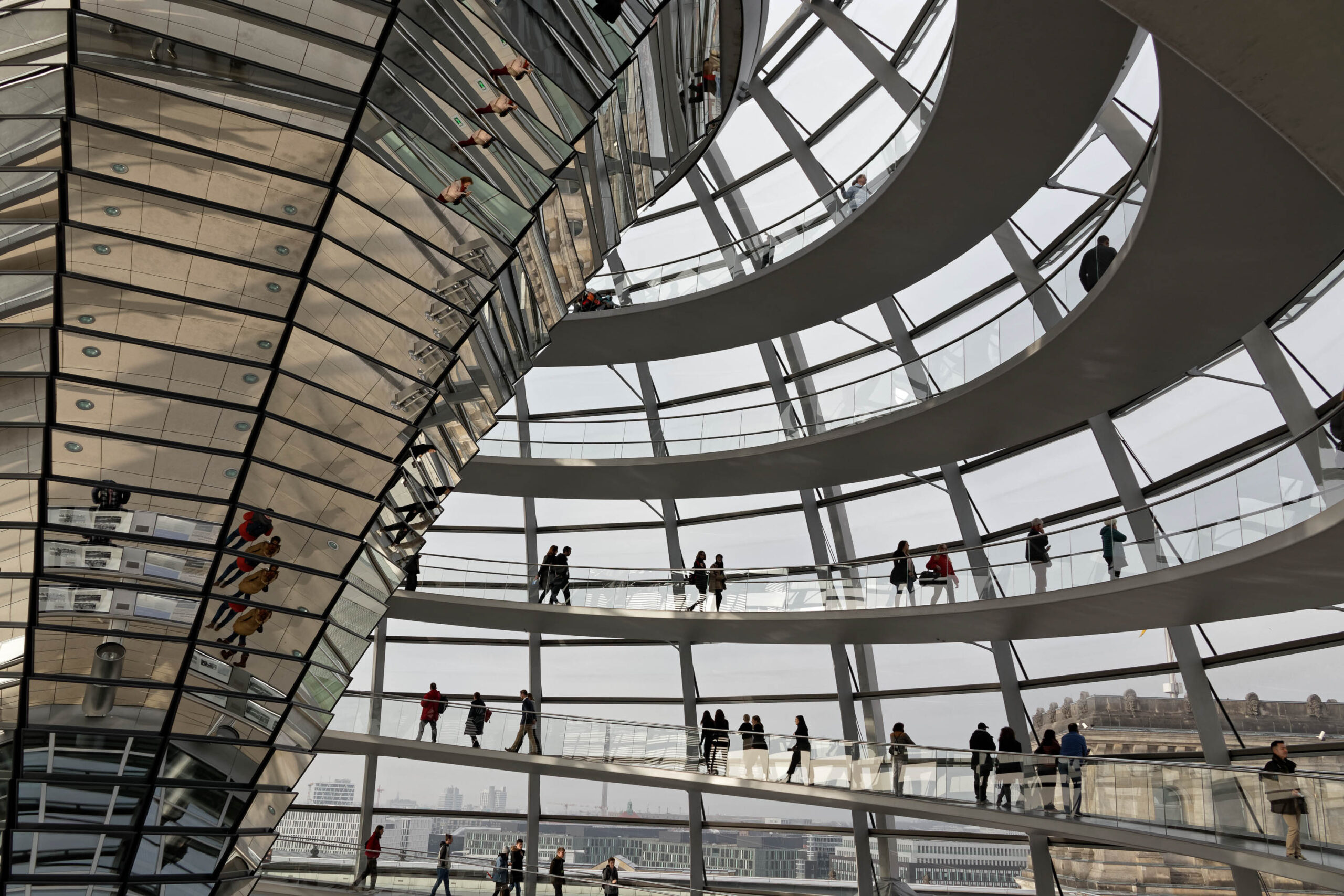 Die Aufnahme zeigt eine Innenansicht der gläsernen Kuppel des Reichstagsgebäudes in Berlin. Die Architektur ist durch die vielen Glasscheiben und Stahlträger geprägt. Im Hintergrund ist die Berliner Skyline zu sehen. Mehrere Personen befinden sich auf den verschiedenen Ebenen der Kuppel und blicken nach draußen. Das Licht fällt durch die Kuppel und erzeugt interessante Licht- und Schatteneffekte.
