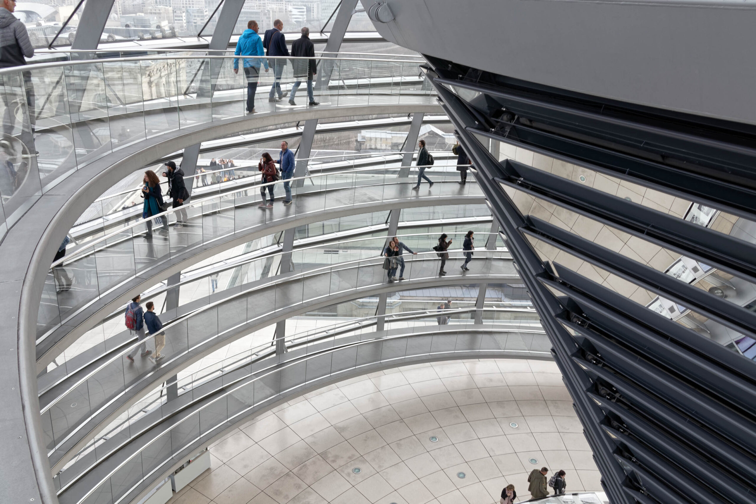 Die Fotografie zeigt eine Innenansicht des Reichstagsgebäudes in Berlin. Die Perspektive ist von oben, wodurch die spiralförmigen Galerieräume und die darunterliegende Kuppel sichtbar werden. Mehrere Personen bewegen sich auf den Galerieräumen, einige stehen, andere gehen. Die Stahlkonstruktion, die die Galerieräume umgibt, ist deutlich erkennbar und lässt einen Blick auf den Himmel und die Stadt durch die Glasflächen zu. Die Kuppel im Zentrum des Bildes ist teilweise durch die Stahlkonstruktion verdeckt. Die Beleuchtung ist diffus und lässt die Szene in einem neutralen Licht erscheinen.