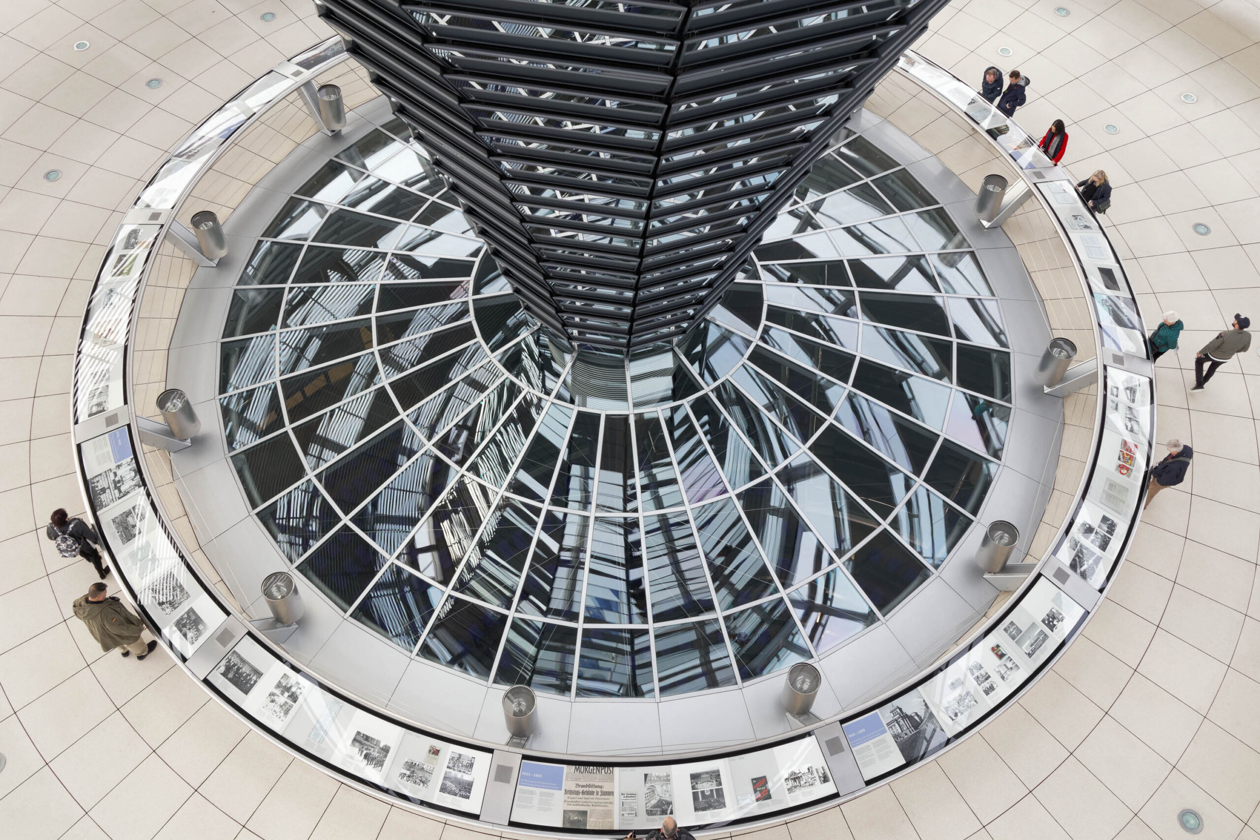 Die Aufnahme zeigt eine Vogelperspektive des Bundestagsgebäudes in Berlin, aufgenommen im März 2019. Das markante Kuppelbauwerk, das das Innere des Gebäudes dominiert, ist von einer Galerie mit fotografischen Dokumentationen umgeben. Menschen bewegen sich im Raum, wobei das Sonnenlicht durch die Glasfassade fällt und interessante Licht- und Schattenmuster erzeugt. Die Architektur ist modern und minimalistisch, mit einer Beton- und Glasbauweise. Die fotografischen Dokumentationen entlang der Kuppel scheinen einen historischen Kontext zu bieten. Die Szene vermittelt eine Atmosphäre der Geschichte, der Politik und der Demokratie.