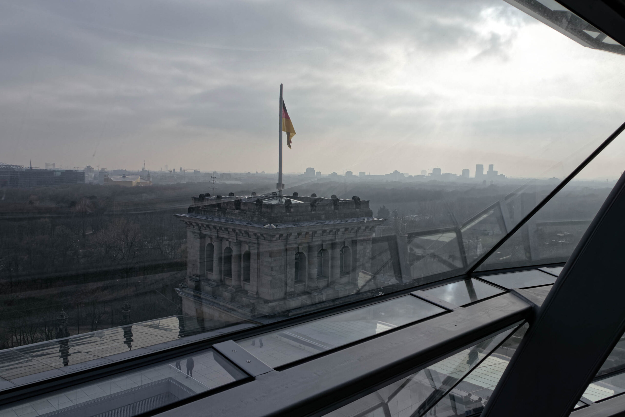 Die Aufnahme zeigt eine Panoramaansicht von Berlin, aufgenommen aus einer erhöhten Position, vermutlich von einem Glasdach oder einer Aussichtsplattform. Im Vordergrund dominiert die Struktur des Glasdachs, das die Sicht auf die Stadt teilweise versperrt. Im Zentrum der Aufnahme befindet sich der Berliner Dom, dessen detaillierte Architektur erkennbar ist. Der Himmel ist bedeckt und die Sichtweite ist aufgrund von Dunst oder Smog eingeschränkt, was die Stadt in einen leicht verschwommenen, grauen Farbton taucht. Im Hintergrund sind weitere Gebäude und die Skyline von Berlin erkennbar, jedoch durch die eingeschränkte Sichtweite weniger deutlich. Die Aufnahme vermittelt eine ruhige, fast melancholische Stimmung.