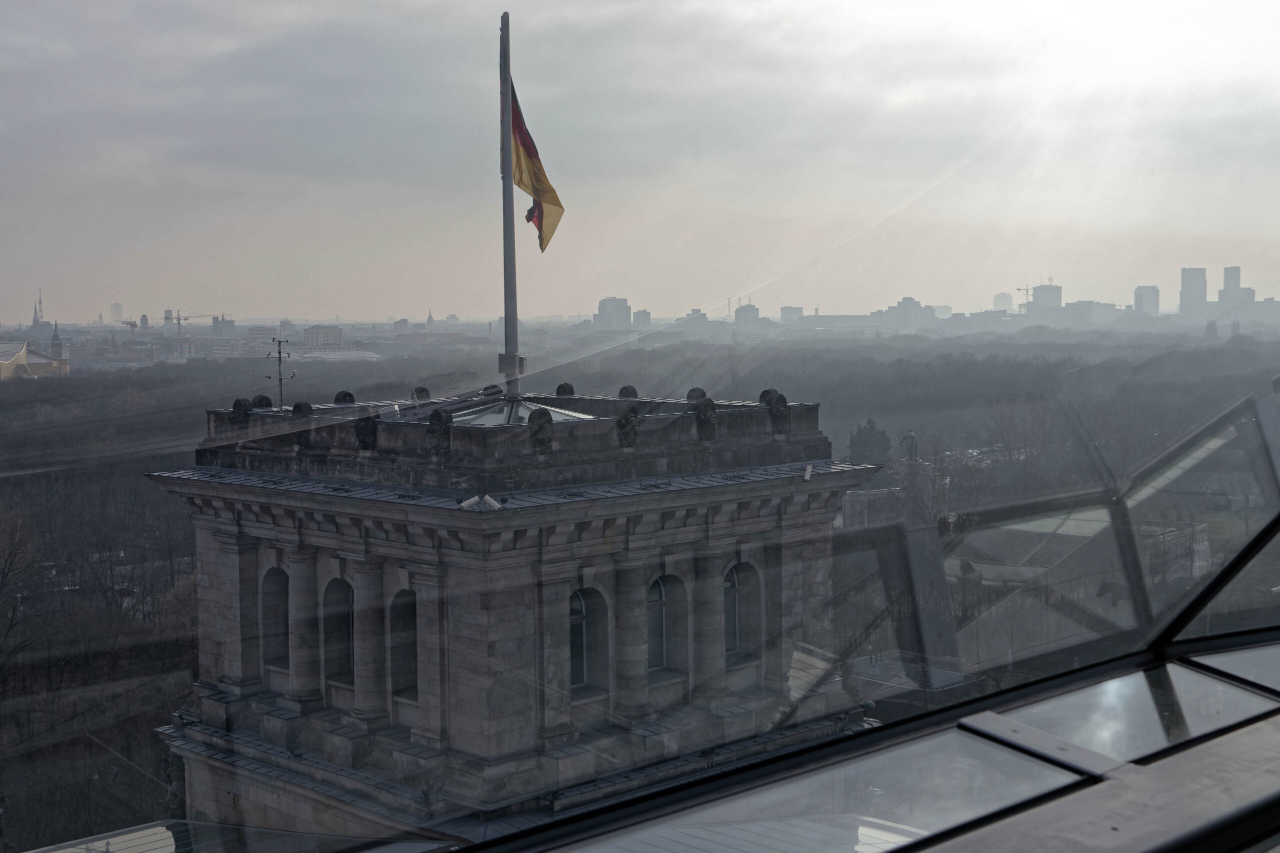 Die Aufnahme zeigt den Berliner Dom bei bedecktem Himmel. Die Perspektive ist von einem erhöhten Standpunkt, vermutlich von einem höheren Gebäude, aufgenommen, was einen weiten Blick über die Stadt ermöglicht. Die Fassade des Doms ist detailliert erkennbar, mit den charakteristischen Kuppeln und Türmen. Im Hintergrund ist die Berliner Skyline mit verschiedenen Gebäuden und Strukturen sichtbar. Der Himmel ist grau und trüb, was eine düstere und melancholische Stimmung erzeugt. Die Aufnahme ist in einem Sepia-Ton gehalten, was den historischen Charakter des Doms und der Stadt unterstreicht. Die Aufnahme ist vermutlich im März 2019 aufgenommen worden.