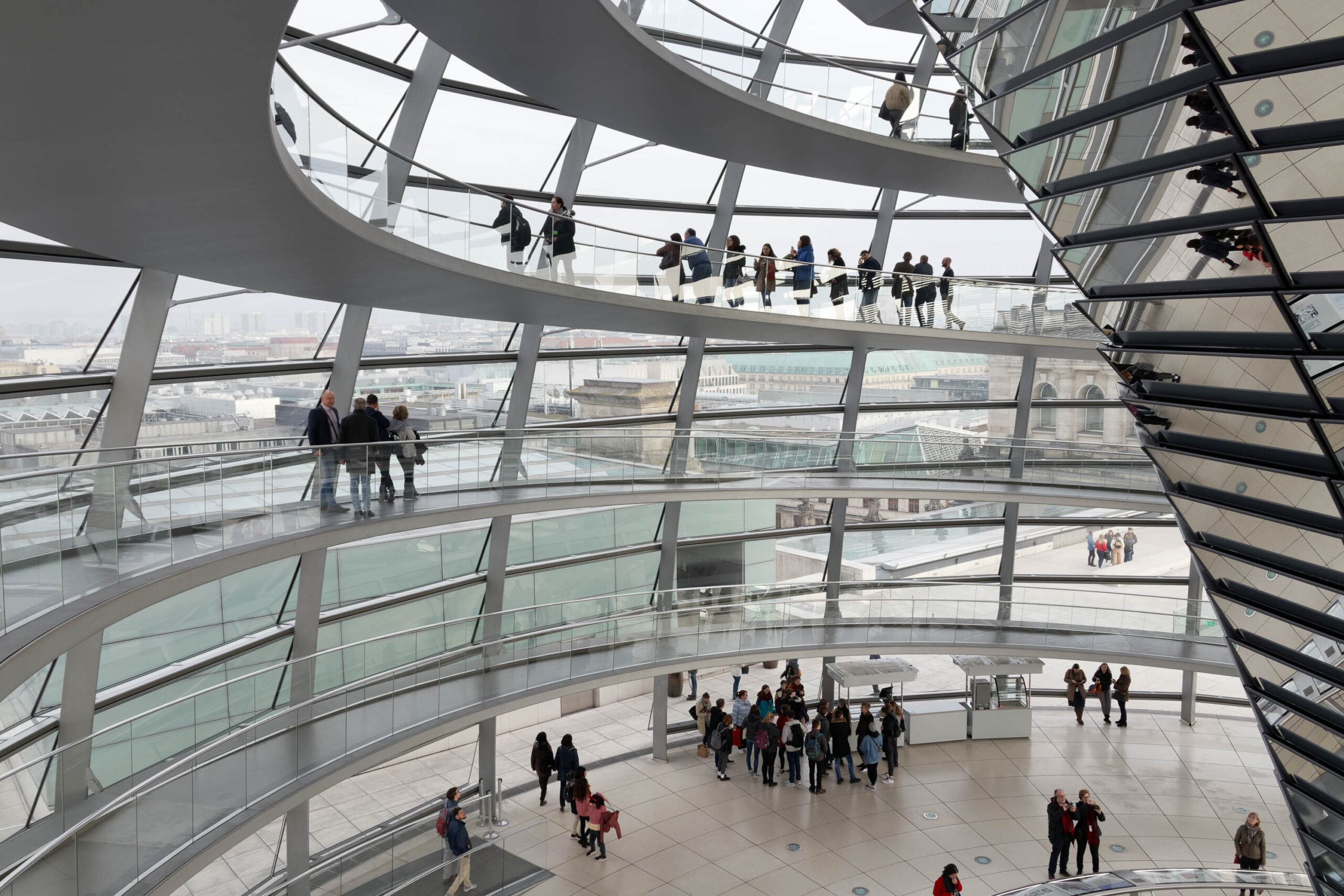 Die Fotografie zeigt eine Innenansicht der Kuppel des Reichstagsgebäudes in Berlin. Die Kuppel ist eine Stahl- und Glaskonstruktion, die einen Panoramablick auf die Stadt ermöglicht. Mehrere Personen befinden sich auf den verschiedenen Ebenen der Kuppel, einige stehen, andere sitzen oder gehen. Die Beleuchtung ist diffus aufgrund des bewölkten Wetters. Die Architektur ist modern und futuristisch, mit geschwungenen Formen und großen Glasflächen. Die Perspektive ist von oben, was die Weite und Höhe der Kuppel betont.