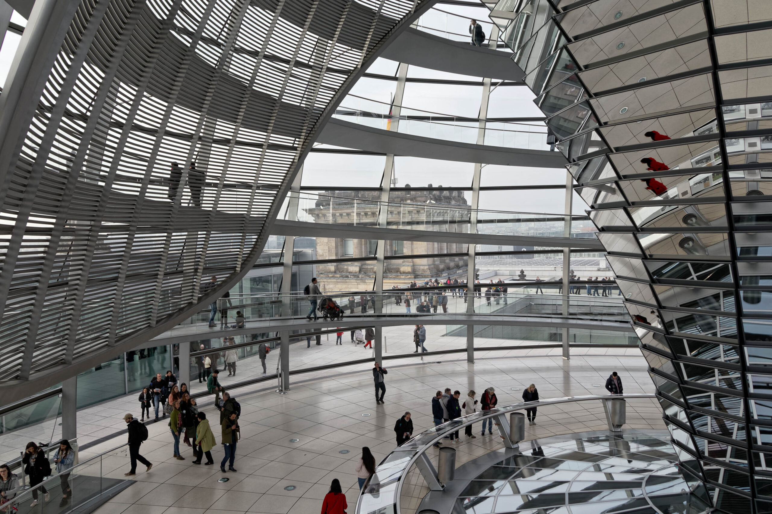 Die Aufnahme zeigt eine Innenansicht der gläsernen Kuppel des Reichstagsgebäudes in Berlin. Die Kuppel besteht aus Stahl- und Glasstrukturen, die einen Panoramablick auf die Stadt ermöglichen. Im Vordergrund befinden sich Besucher, die sich auf den verschiedenen Ebenen der Kuppel aufhalten. Im Hintergrund ist die Berliner Skyline mit verschiedenen Gebäuden und dem Himmel zu sehen. Das Licht ist diffus und erzeugt eine ruhige Atmosphäre.