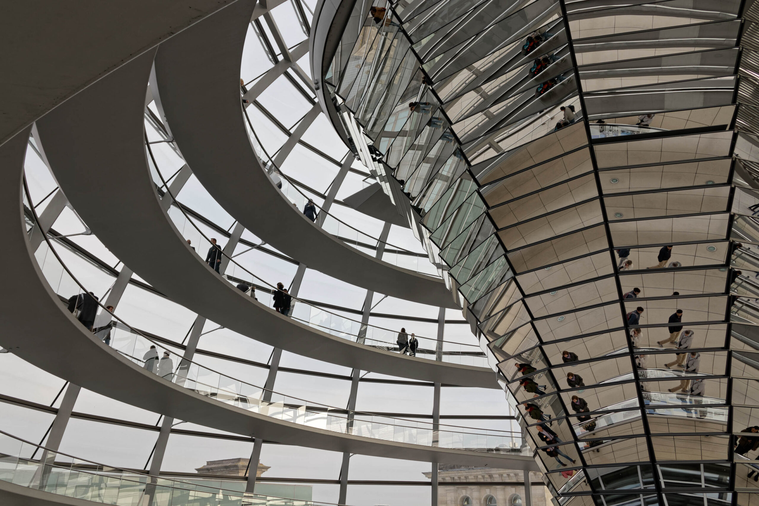 Die Aufnahme zeigt eine detaillierte Innenansicht der Kuppel des Reichstagsgebäudes in Berlin. Die Architektur ist durch die komplexen, spiralförmigen Glaspaneele dominiert, die einen transparenten Blick nach oben ermöglichen. Mehrere Personen sind auf den Galerien zu sehen, die sich in der Kuppel winden, wobei einige sich bewegen und andere stehen. Das Licht fällt durch die Glaspaneele und erzeugt ein Spiel von Schatten und Reflexionen. Die Gesamtstimmung ist ruhig und kontemplativ, vermittelt durch die weiten, offenen Räume und die subtile Beleuchtung.