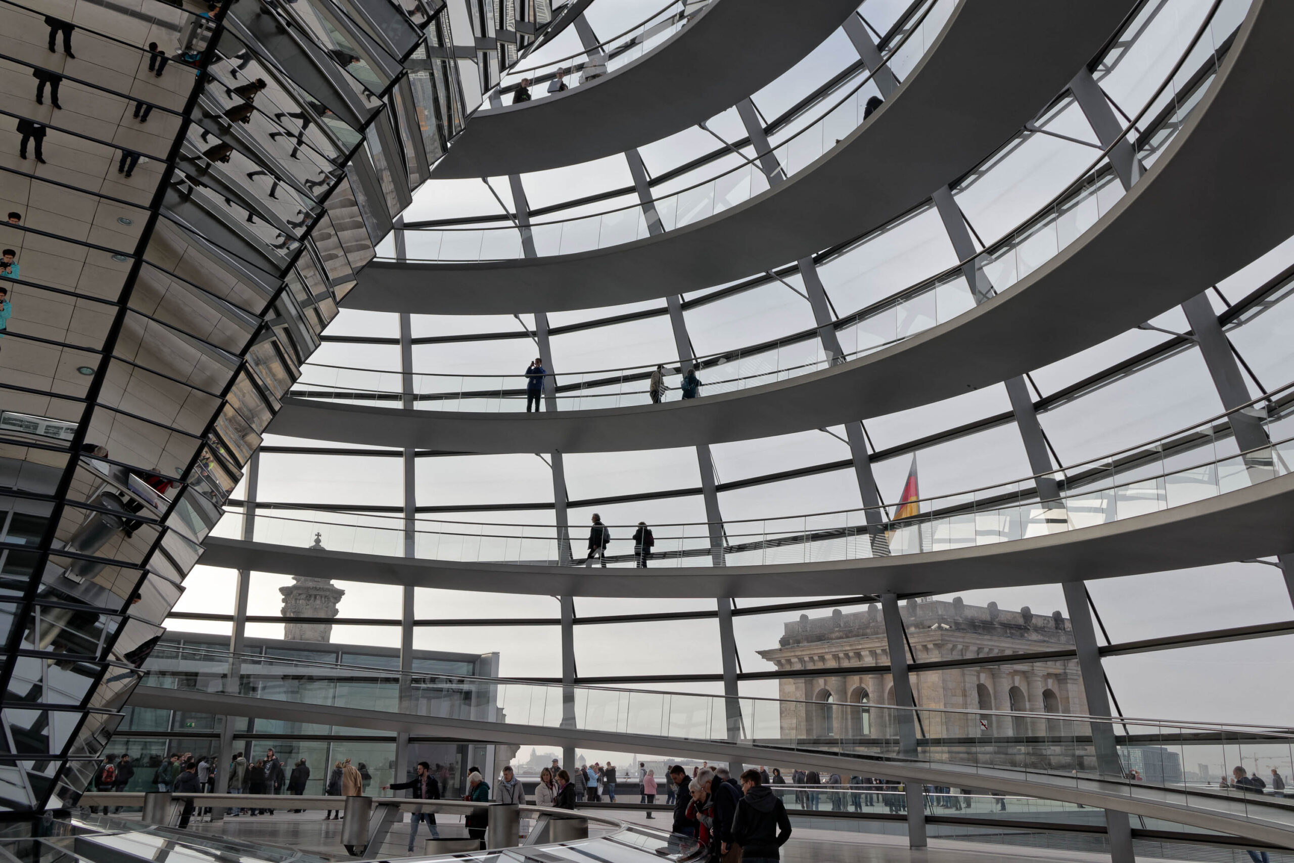 Die Aufnahme zeigt eine Innenansicht der Kuppel des Reichstagsgebäudes in Berlin. Die Kuppel besteht aus einer komplexen Struktur aus Stahl und Glas, die einen Panoramablick auf die Stadt ermöglicht. Mehrere Personen sind auf verschiedenen Ebenen der Kuppel zu sehen, einige blicken nach draußen, andere interagieren miteinander. Im Hintergrund sind Gebäude und der Himmel sichtbar. Die Beleuchtung ist diffus, was eine ruhige und friedliche Atmosphäre schafft.