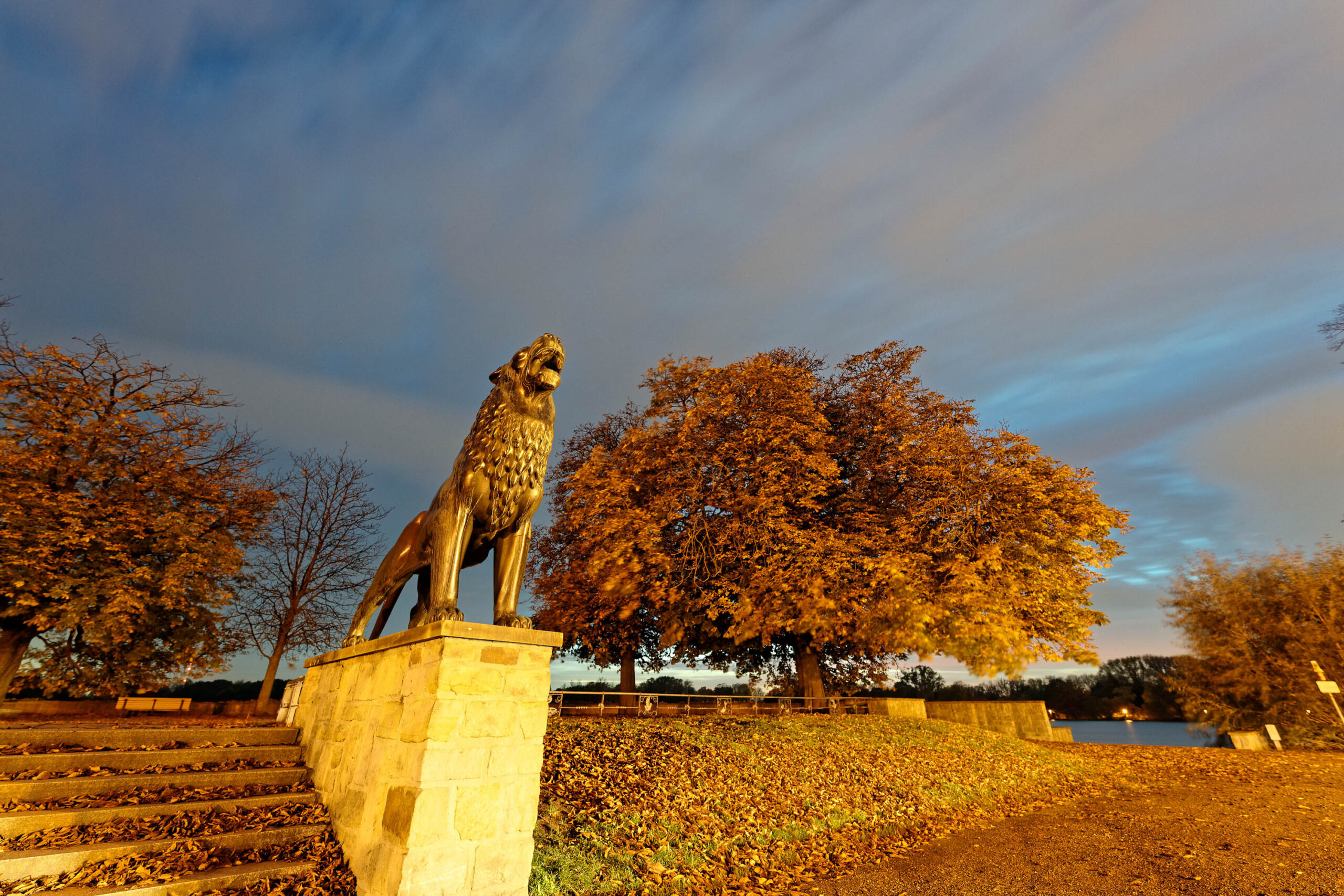 Das Foto zeigt den Löwenbrunnen in Hannover bei Dämmerung. Der Löwenbrunnen ist ein markantes Denkmal und steht auf einem Sockel aus Ziegelsteinen. Der Löwe ist aus Bronze und scheint im Licht zu leuchten. Im Hintergrund ist ein See und Bäume zu sehen, deren Blätter sich in Herbstfarben präsentieren. Die Szene wird von einem blauen Himmel mit einigen Wolken und einem warmen Schein am Horizont beleuchtet. Ein paar Stufen führen zu dem Brunnen hinauf. Die Atmosphäre ist ruhig und friedlich.