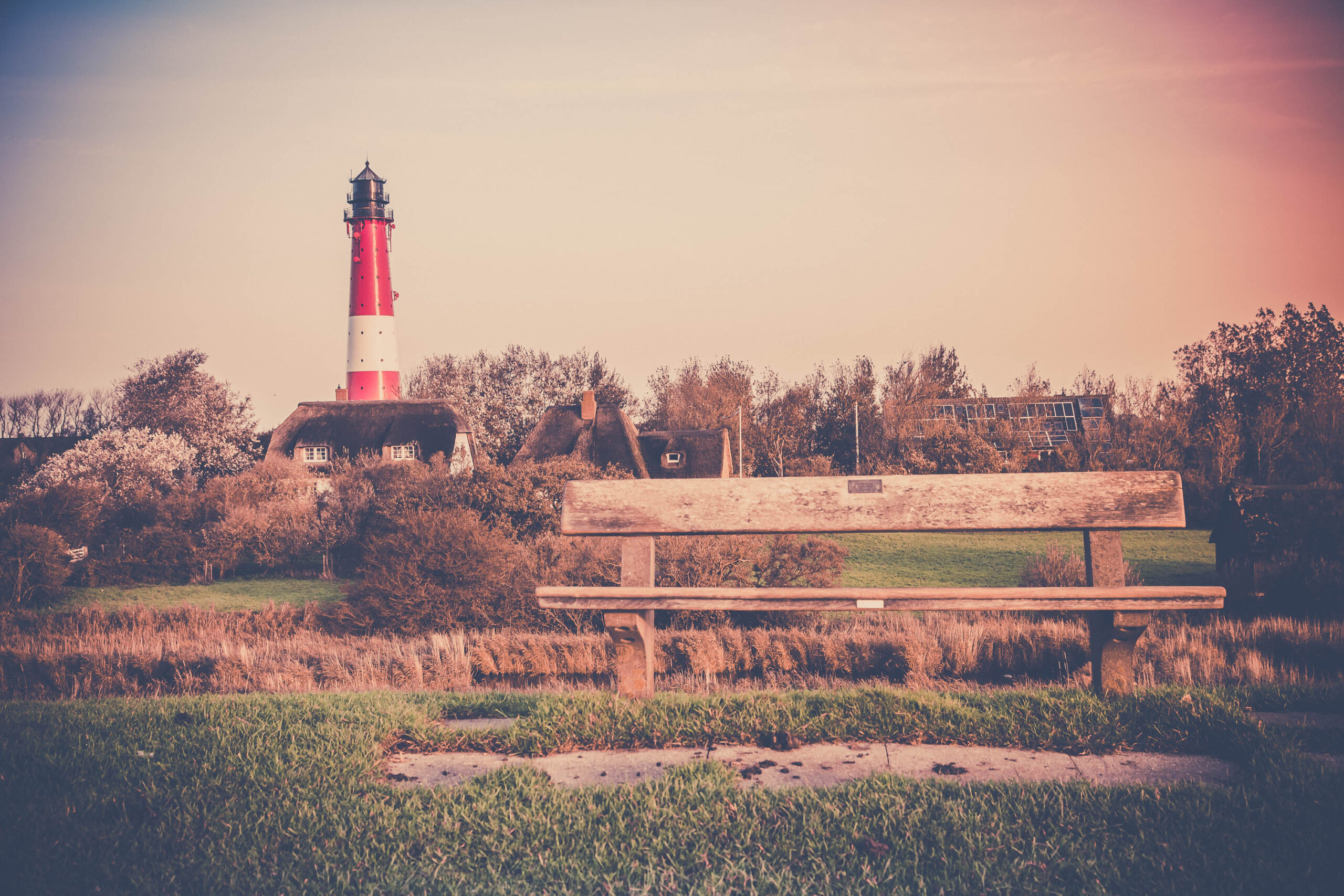 Die Aufnahme zeigt eine idyllische Szene auf der Insel Pellworm im Wattenmeer. Im Vordergrund steht eine einfache Holzbank, die einen Blick auf einen markanten, weiß-rot gestreiften Leuchtturm freigibt. Im Hintergrund sind traditionelle Häuser mit charakteristischen Strohdächern zu erkennen, die in die Landschaft integriert sind. Die Farben sind gedämpft und wirken nostalgisch, was durch die warme Beleuchtung und die leicht verwaschene Optik verstärkt wird. Die Szene vermittelt ein Gefühl von Ruhe und Abgeschiedenheit, typisch für die nordfriesischen Inseln.