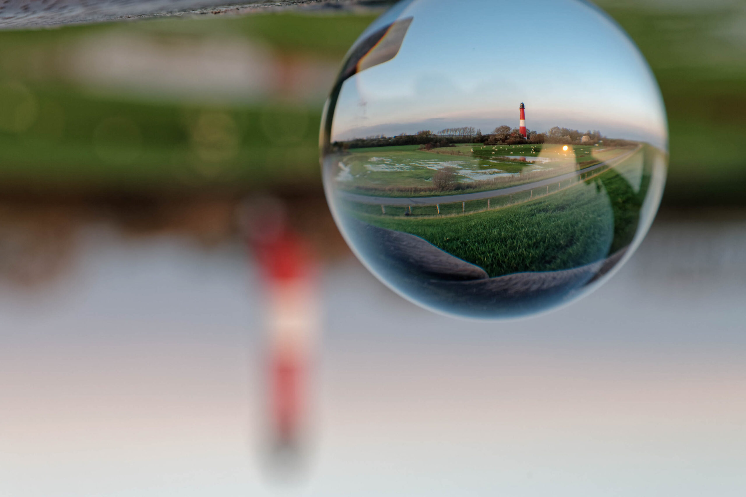 Die Aufnahme zeigt eine ungewöhnliche Perspektive auf eine norddeutsche Landschaft. Im Vordergrund liegt eine Kugel, die die umliegende Landschaft in sich spiegelt. Diese Spiegelung zeigt einen Leuchtturm, der sich in der Ferne erhebt, umgeben von flachem Grünland und einem bewölkten Himmel. Die Kugel liegt auf einer Holzkonstruktion, die wiederum auf einer grasbewachsenen Fläche ruht. Die Farben sind gedämpft und wirken melancholisch. Die Aufnahme erzeugt eine surreale und verträumte Atmosphäre.