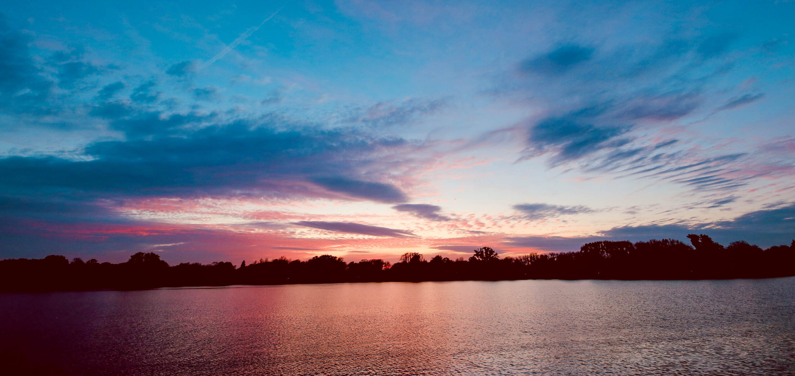 Die Fotografie zeigt einen atemberaubenden Sonnenuntergang über einem See in der Nähe von Hannover. Der Himmel ist in leuchtenden Farben von Rosa, Violett und Orange gehalten, die sich in der ruhigen Wasseroberfläche spiegeln. Ein dunkler Baumhorizont bildet den Hintergrund, wodurch die Farben des Himmels noch intensiver wirken. Die Szene vermittelt eine friedliche und ruhige Atmosphäre.