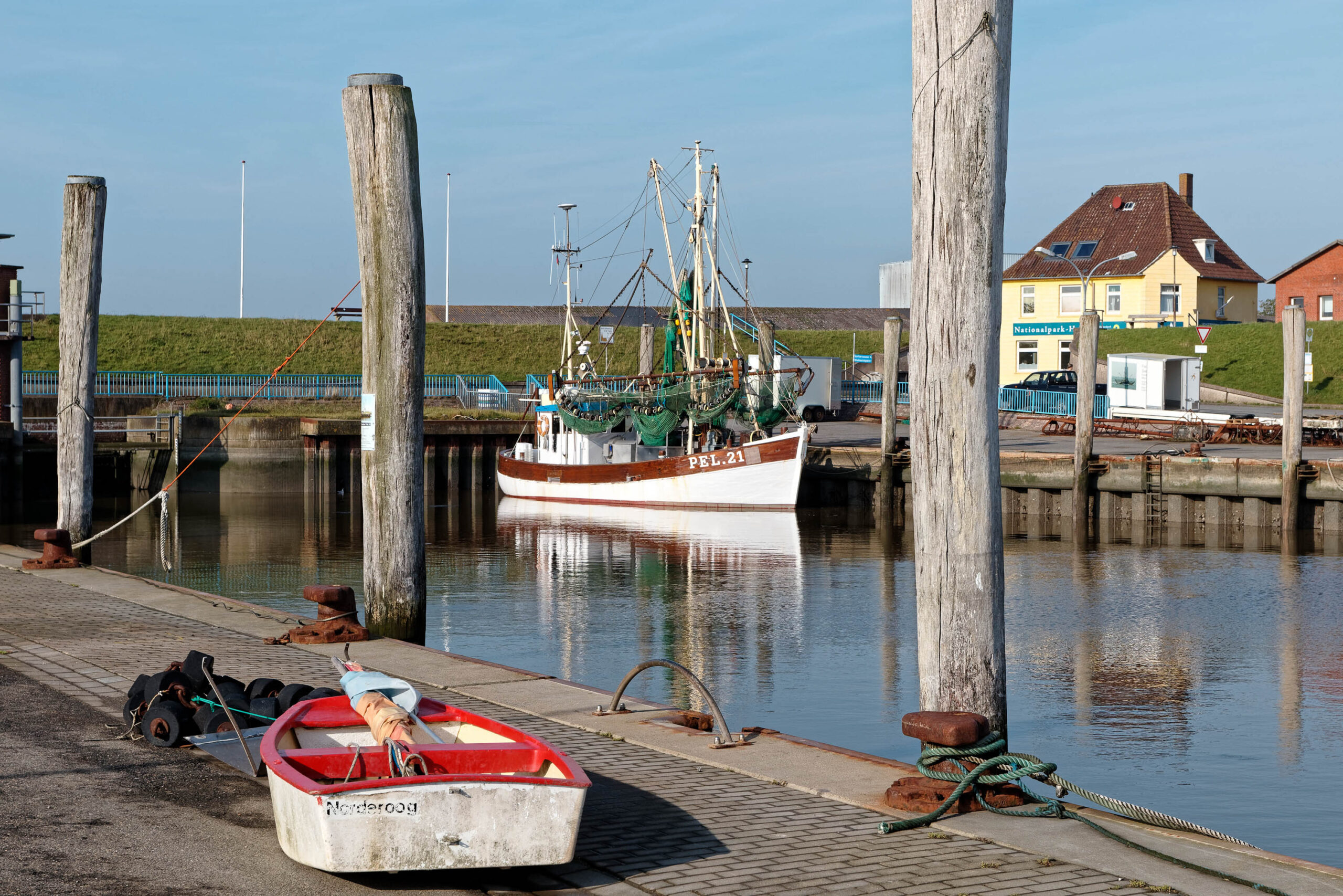 Das Bild zeigt einen Hafen in Pellworm, Nordsee, im Oktober 2019. Im Vordergrund liegt ein kleines, rotes Ruderboot, das teilweise mit Ausrüstung beladen ist. Im Hintergrund befindet sich ein Fischerboot mit dem Schriftzug „PEL 21“ auf dem Rumpf.  Hinter dem Fischerboot sind typische Häuser mit roten Ziegeldächern und gelben Fassaden zu sehen, die für die Architektur von Pellworm charakteristisch sind. Die Szene wird von hölzernen Pfählen und einer Hafenpromenade eingerahmt. Das Wasser spiegelt die Szene wider und erzeugt eine ruhige Atmosphäre.