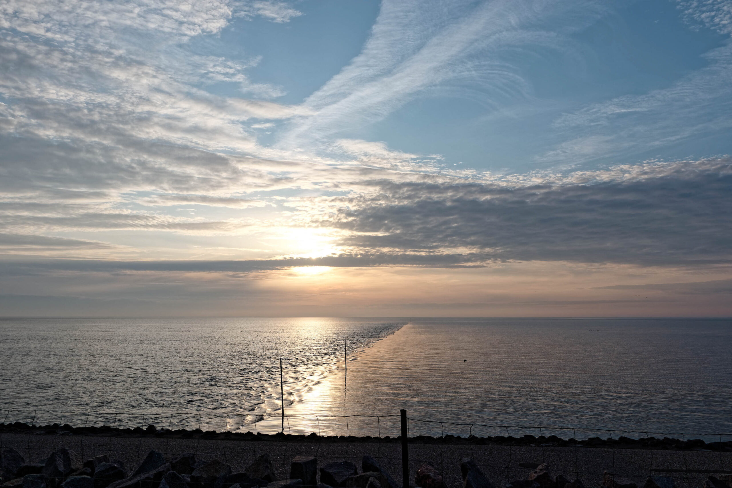 Das Bild zeigt einen Sonnenaufgang über dem Meer, aufgenommen auf der Insel Pellworm im Oktober 2019. Der Himmel ist von einem sanften Blau bedeckt, das sich in Richtung des Horizonts in ein warmes Orange übergeht. Zahlreiche, feine Wolken ziehen über den Himmel und reflektieren das Licht der aufgehenden Sonne. Das Meer ist ruhig und spiegelt das Licht der Sonne und des Himmels wider, wodurch ein schimmernder Effekt entsteht. Im Vordergrund befindet sich ein dunkler Strand, der durch einen Zaun begrenzt ist. Die Silhouette des Strandes und des Zauns bildet einen starken Kontrast zum hellen Himmel und dem Meer. Die Szene vermittelt eine friedliche und ruhige Atmosphäre.