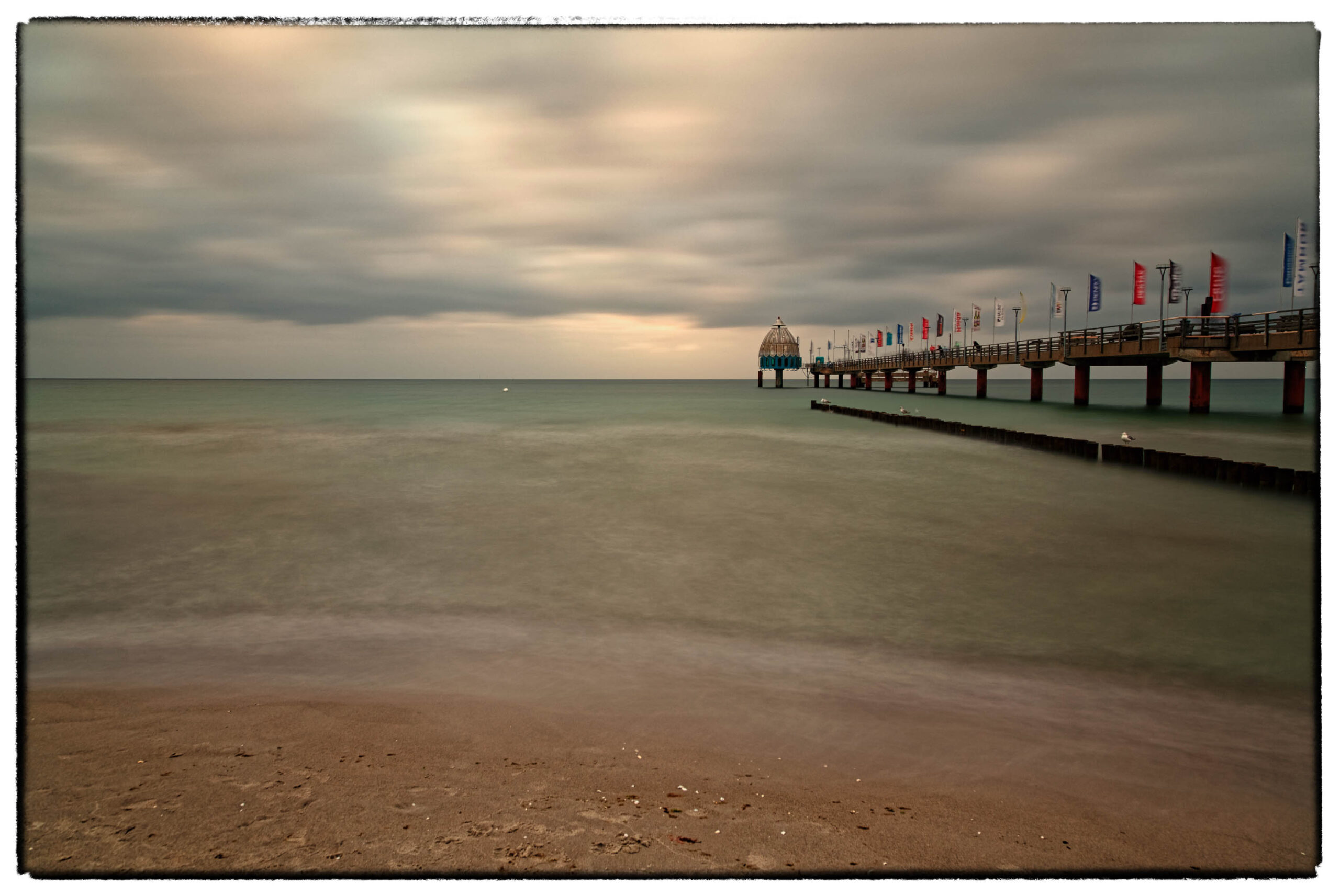 Das Bild zeigt einen Strand in Zingst an der Ostsee. Im Vordergrund liegt ein Sandstrand, der in den ruhigen, trüben Meereswasser übergeht. Ein hölzerner Steg, vermutlich ein Seebrücke, erstreckt sich weit in das Meer hinein. Auf dem Steg befinden sich Personen, die sich in Bewegung befinden, was durch die Bewegungsunschärfe angedeutet wird. Der Himmel ist stark bewölkt, was eine düstere und melancholische Stimmung erzeugt. Die Farben sind gedämpft und wirken fast monochrom, was die Atmosphäre des Bildes verstärkt. Die Aufnahme wurde im Juli 2019 aufgenommen.